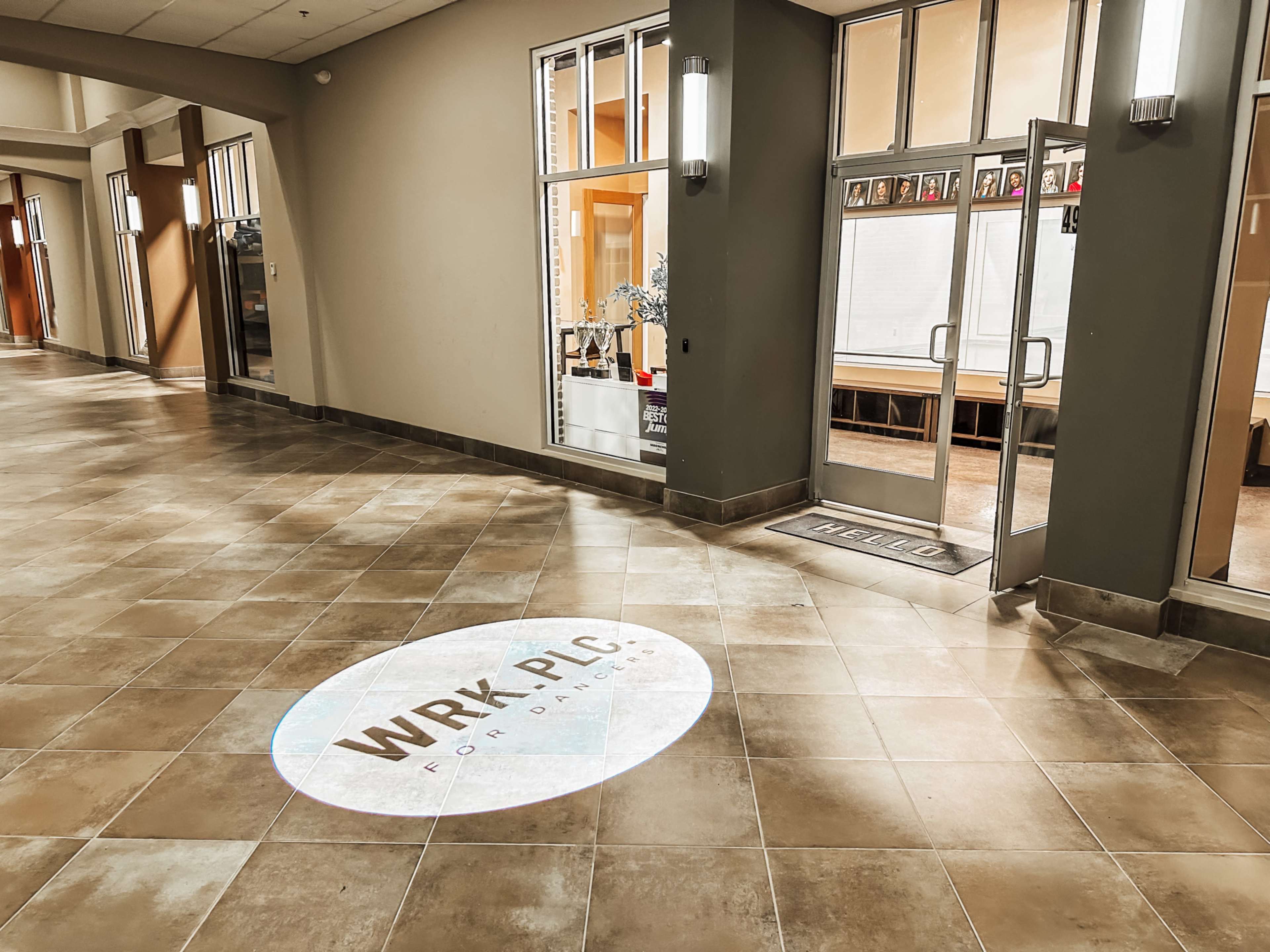 The image shows a hallway with tiled flooring and large windows leading to a room displaying the text "WRK-PLC" on the floor.