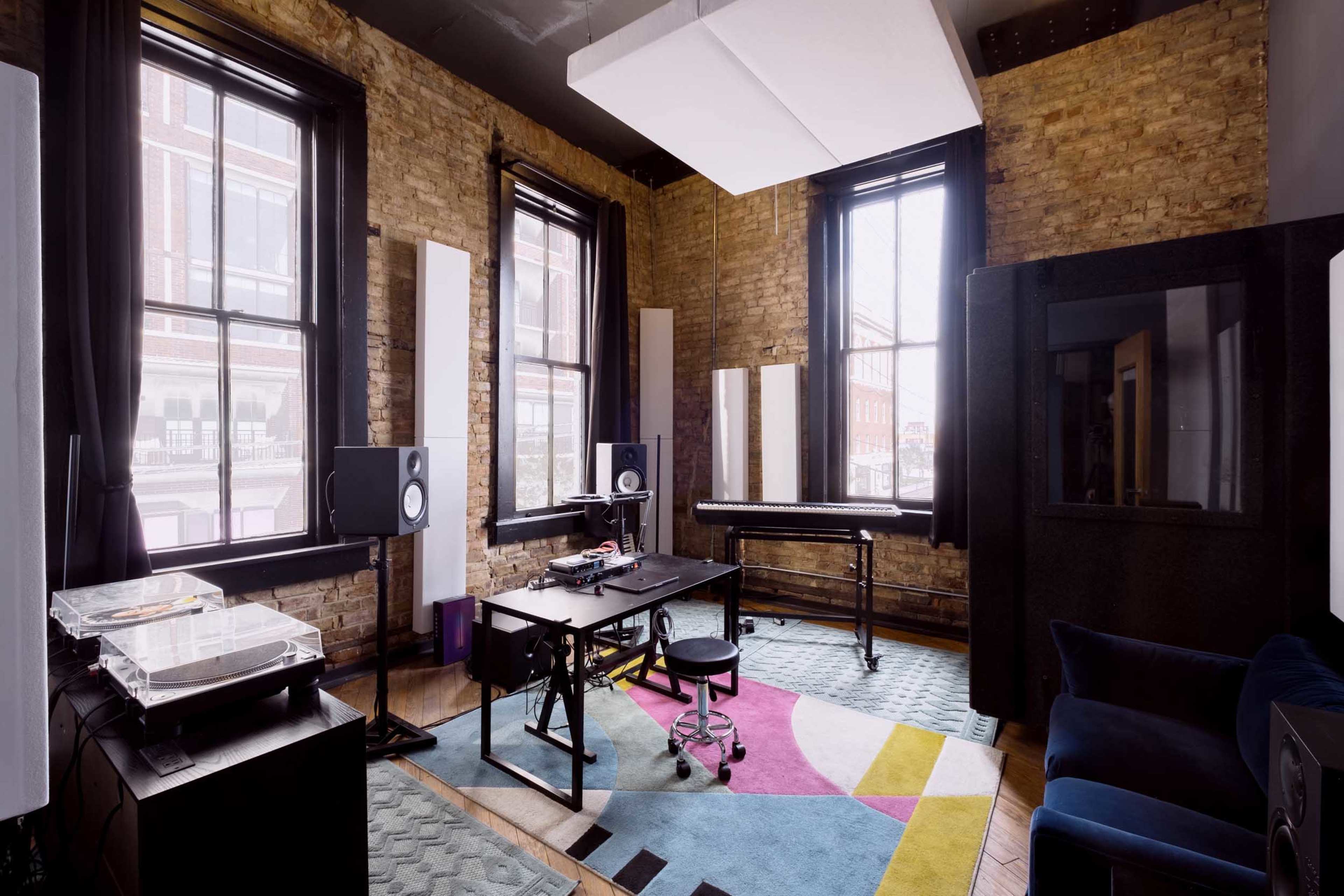 The image shows a music studio with large windows, brick walls, a desk with equipment, and a colorful rug on the floor.