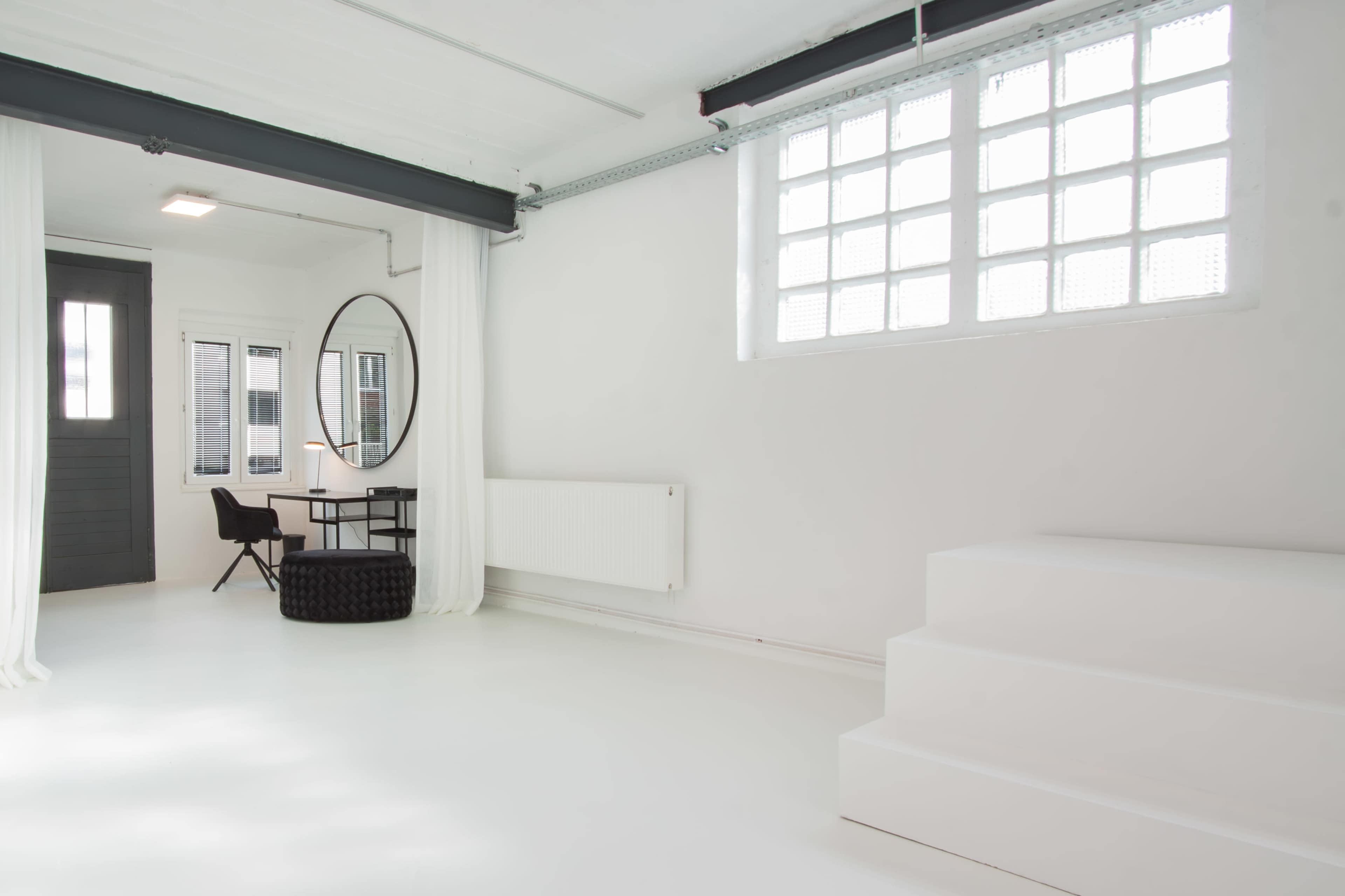 A minimalist interior space with white walls, a circular mirror, a desk and chair, and a small staircase leading to a raised section.