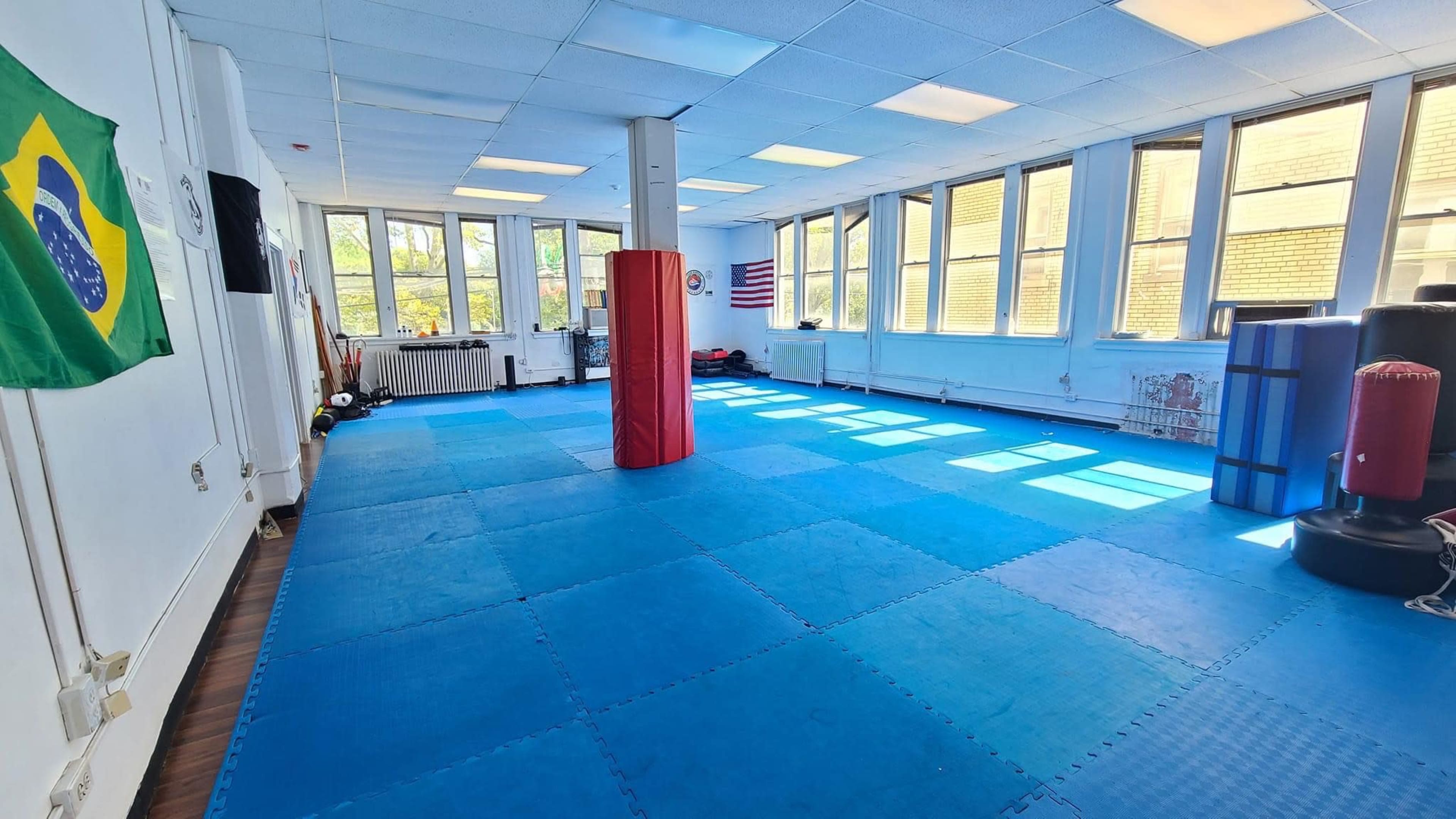 The image shows a spacious martial arts studio with blue floor mats, various training equipment, and large windows allowing natural light to enter.