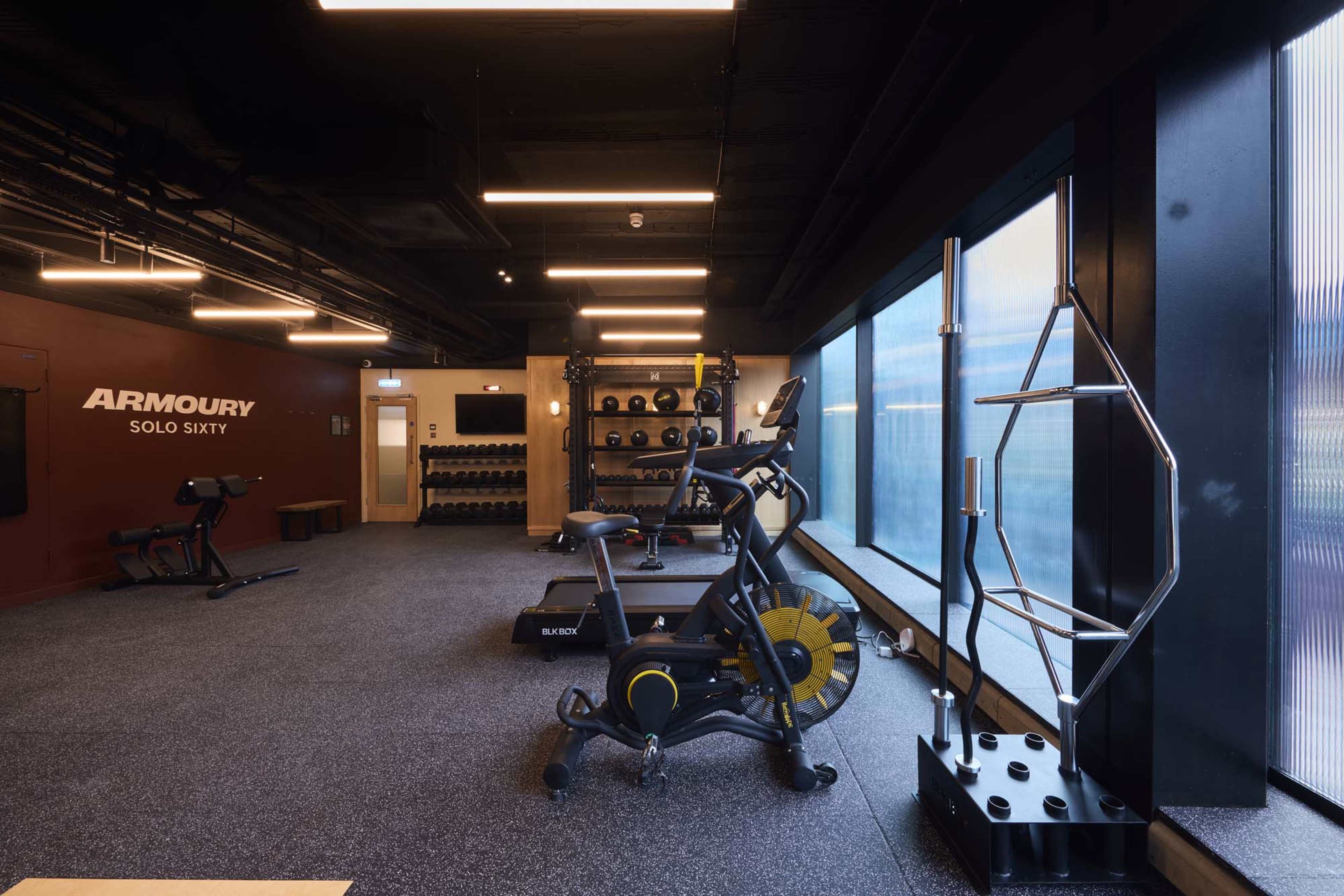 The image depicts a modern gym space featuring exercise equipment such as a stationary bike, weights, and a stair climber, with large windows allowing natural light.