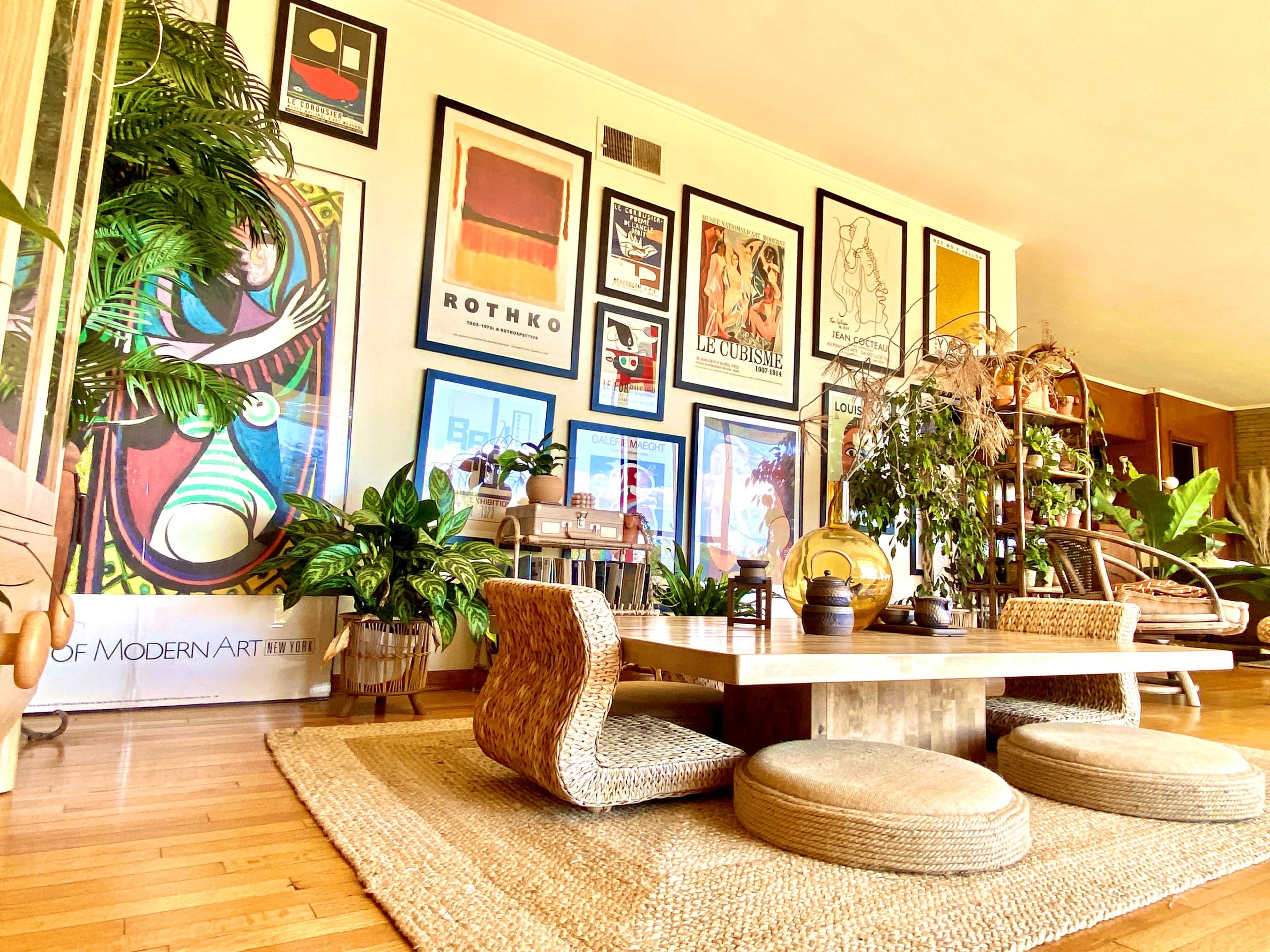 The image depicts a modern living room featuring a large wall adorned with colorful art prints, a low wooden table surrounded by round woven seats, and various indoor plants throughout the space.