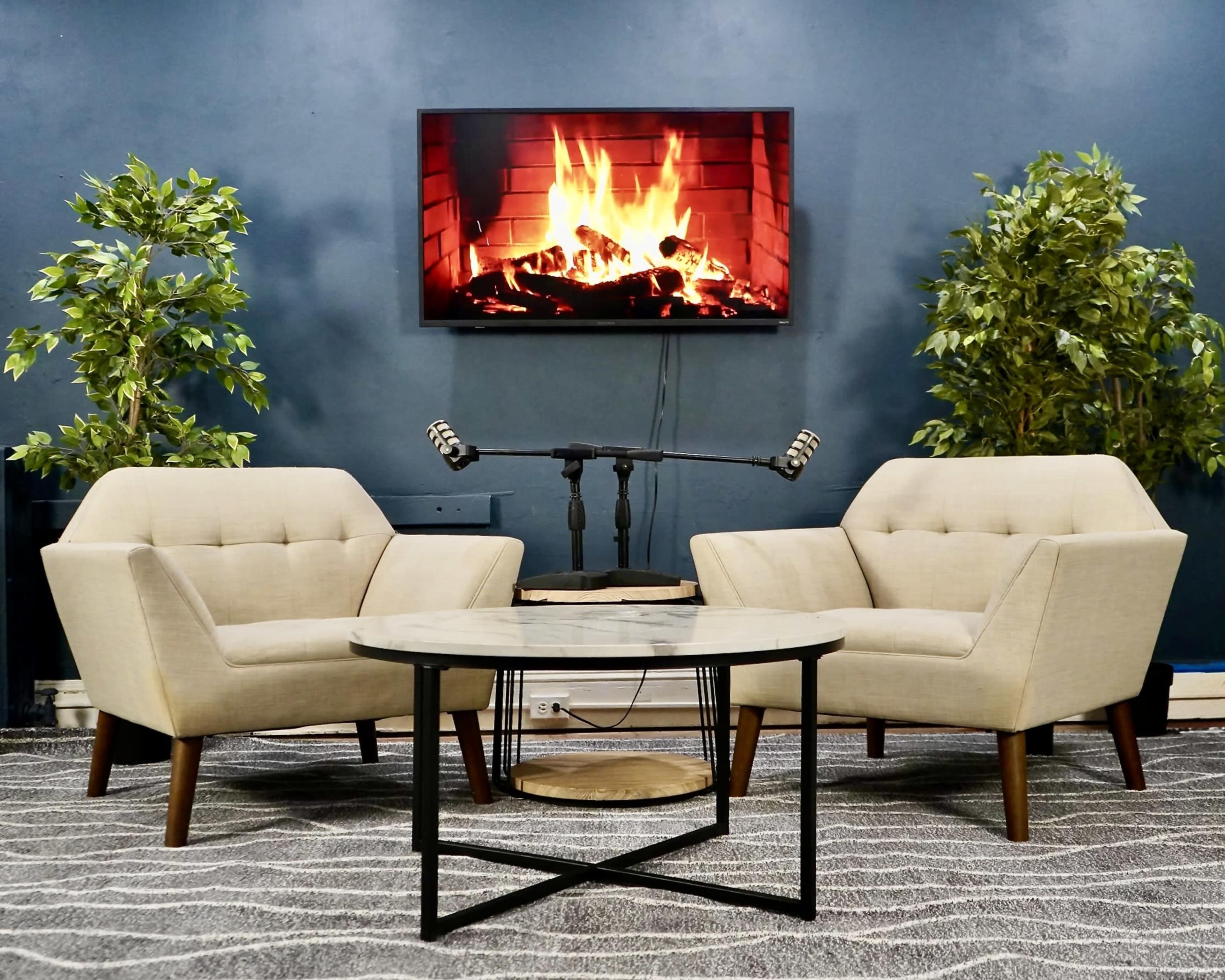 The image shows a cozy seating area with two light-colored chairs facing a circular table, set against a blue wall featuring a television displaying a fireplace.