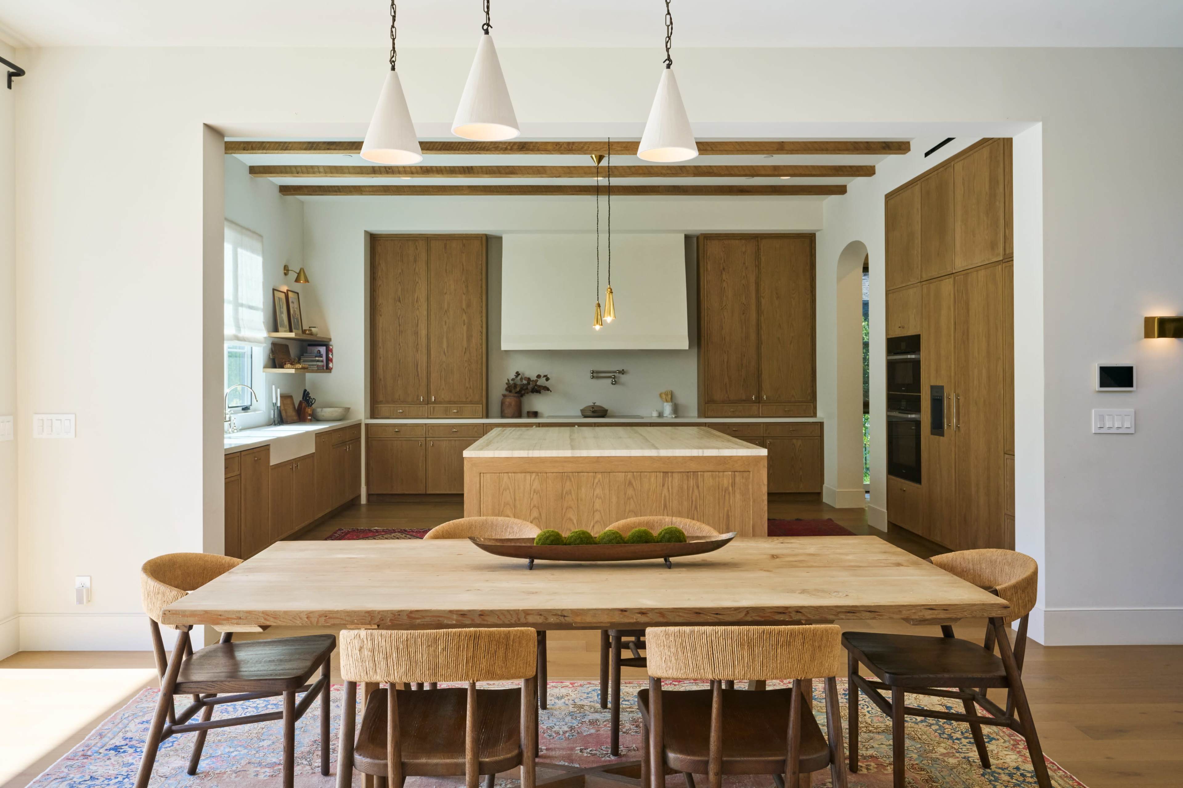 The image shows a modern kitchen and dining area featuring wooden cabinetry, a large central island, and a wooden dining table surrounded by various chairs.