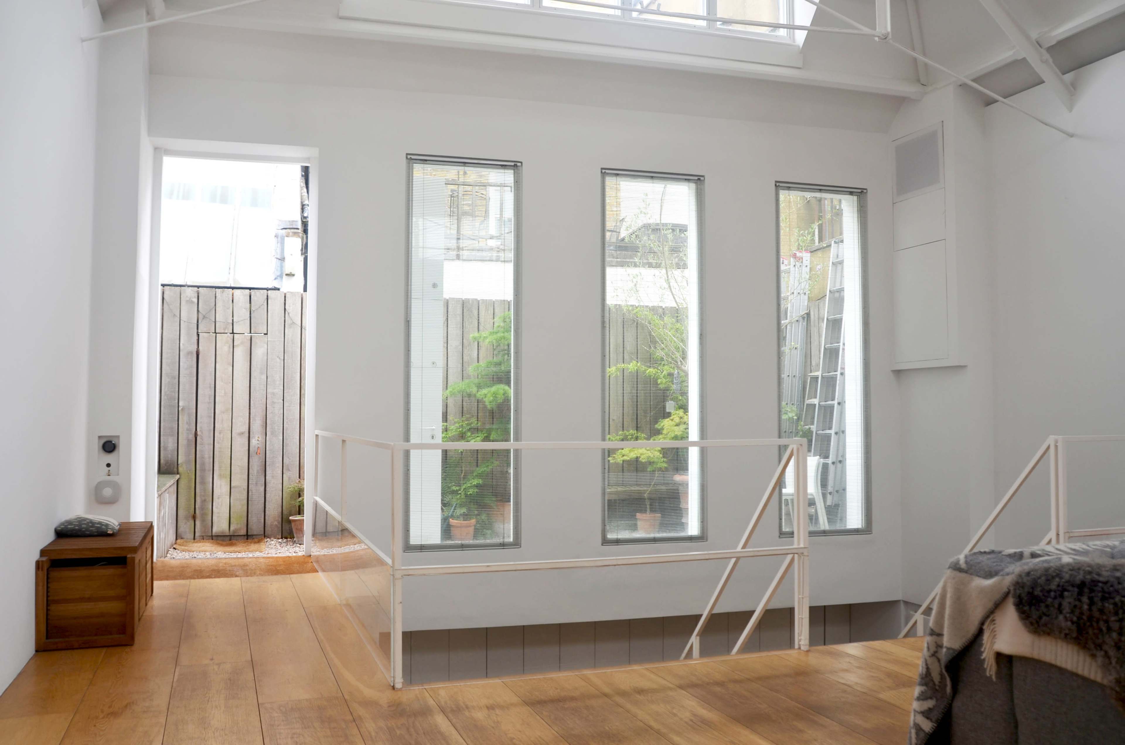 The image shows a bright, modern interior space with large windows overlooking a small outdoor area featuring plants and wooden fencing.