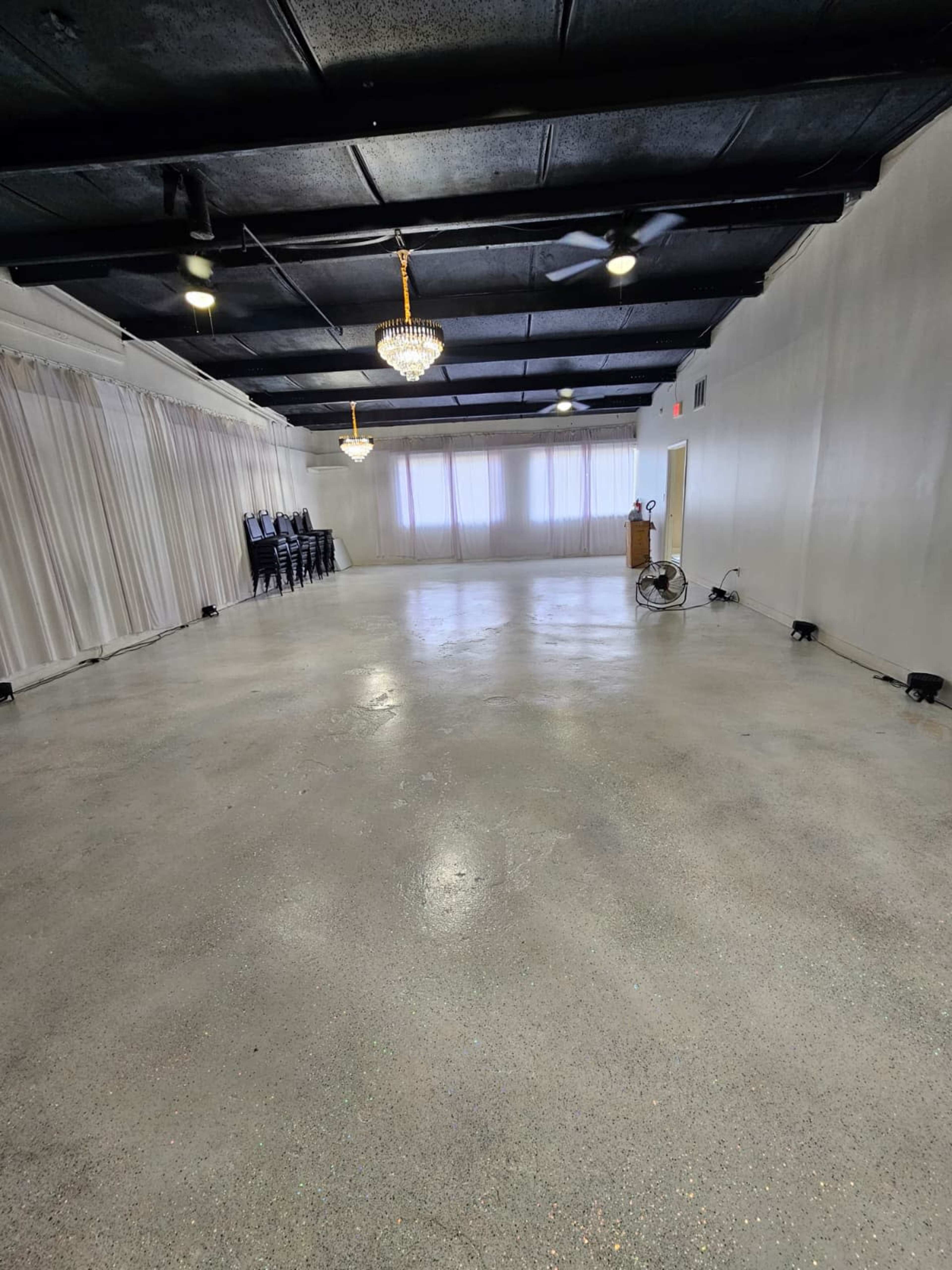 The image shows a spacious, empty room with a polished concrete floor, white walls, and a row of chairs along one side.