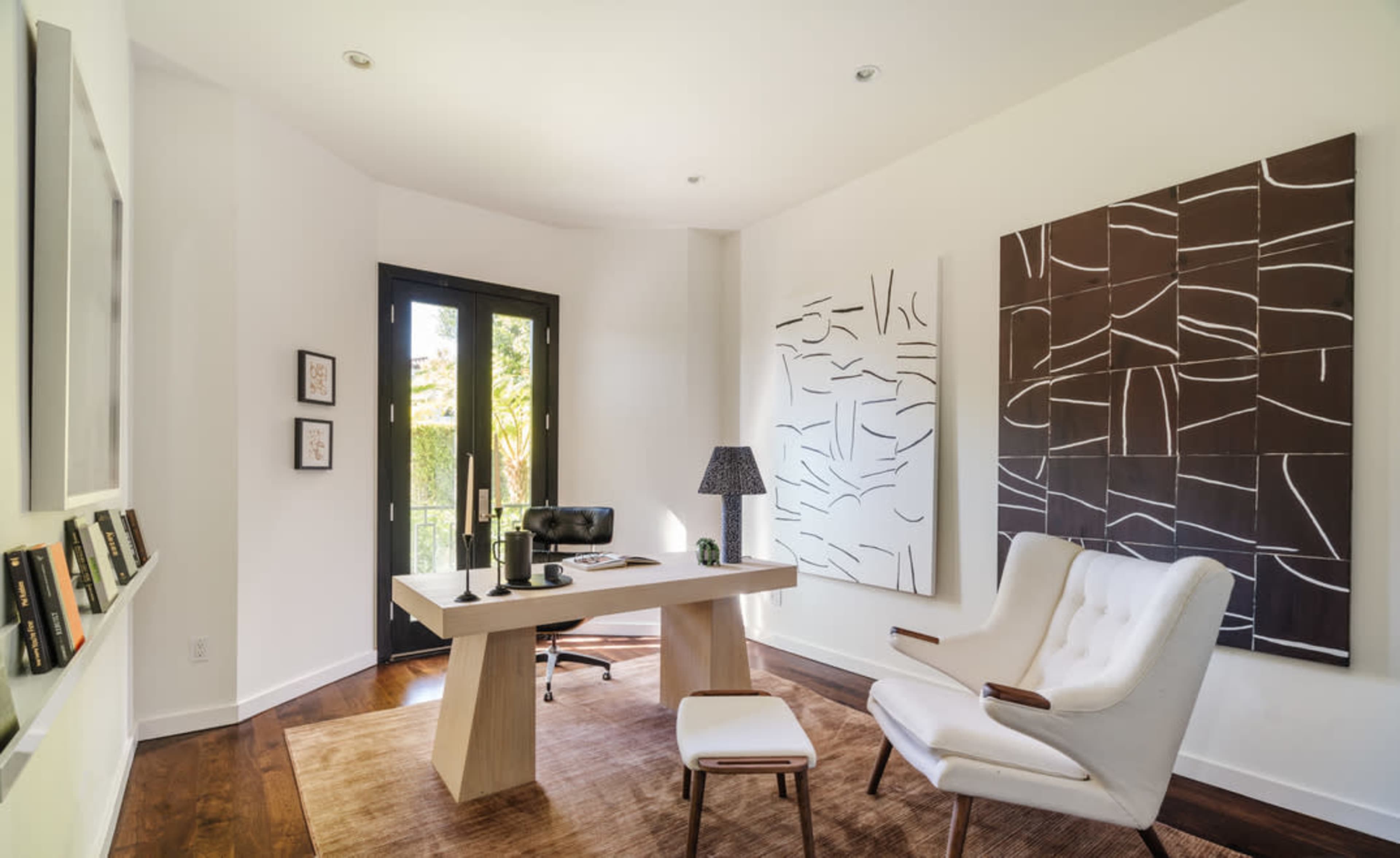 A modern office features a light wood desk, a white armchair, and abstract wall art, with large windows allowing natural light.
