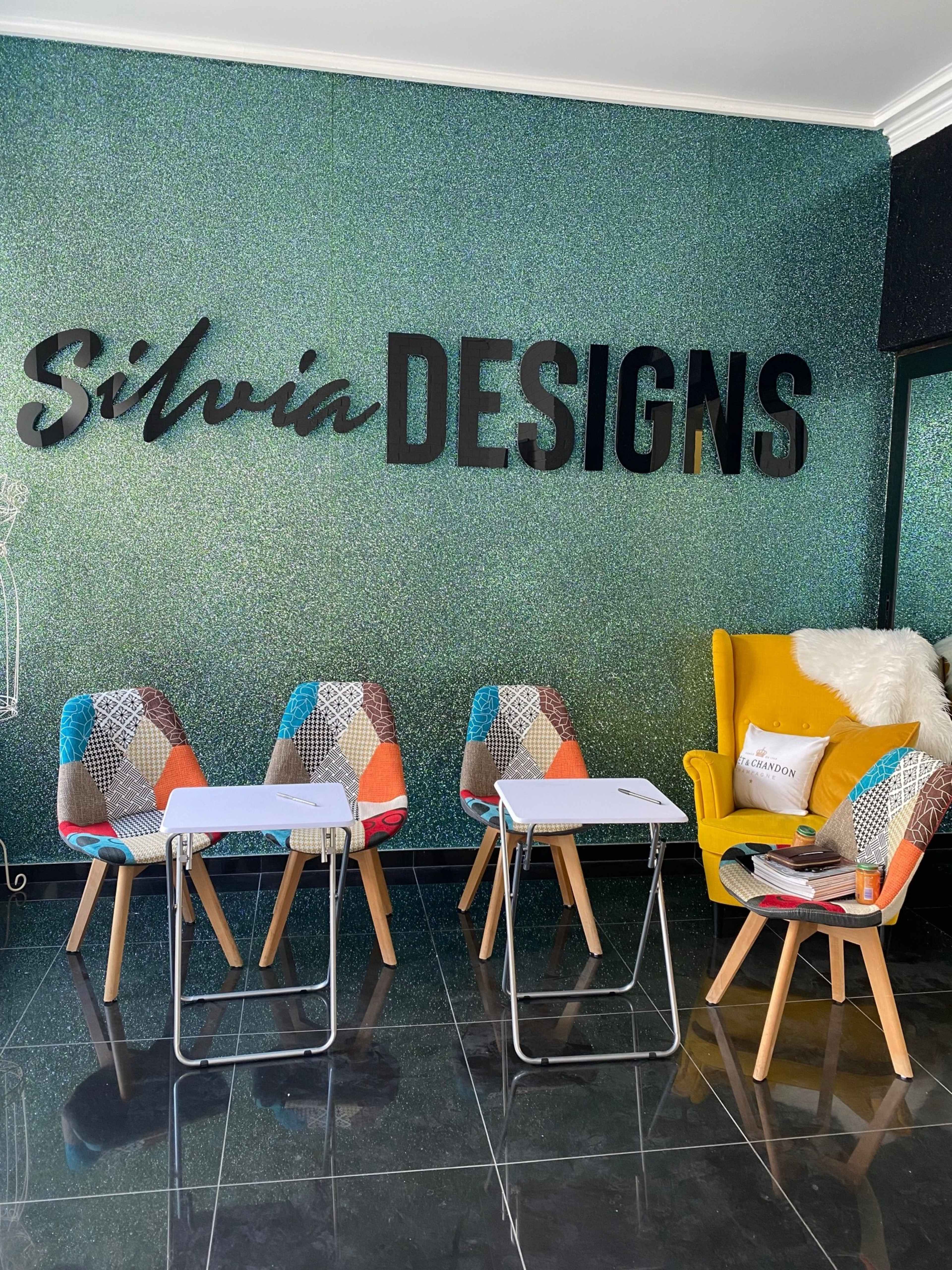 A modern interior with colorful chairs and a yellow armchair, featuring a wall sign that reads "Silvia DESIGNS" against a sparkling green background.