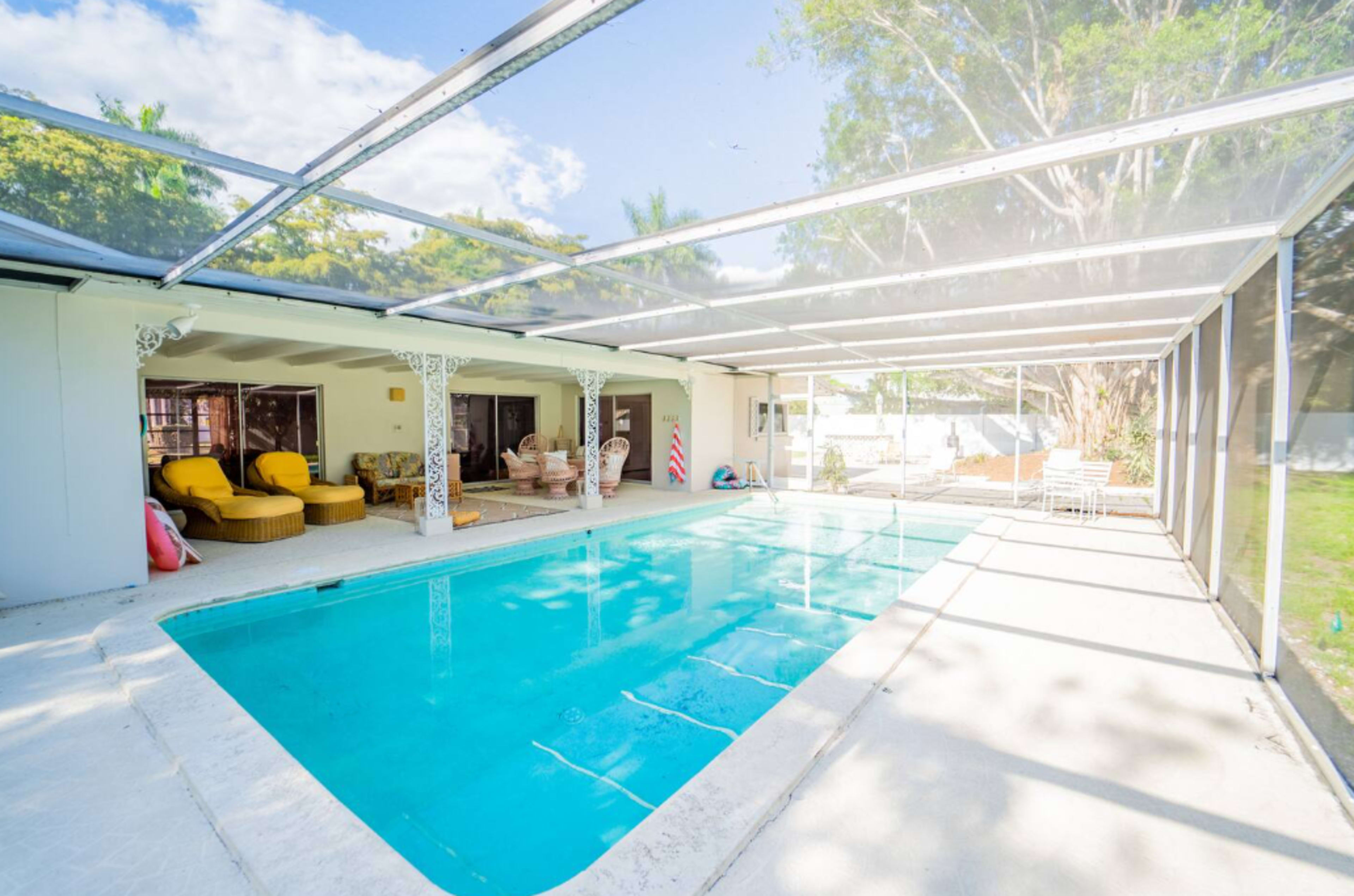 A spacious screened-in pool area features a clear blue pool, yellow lounge chairs, and a backdrop of greenery and sunlight.