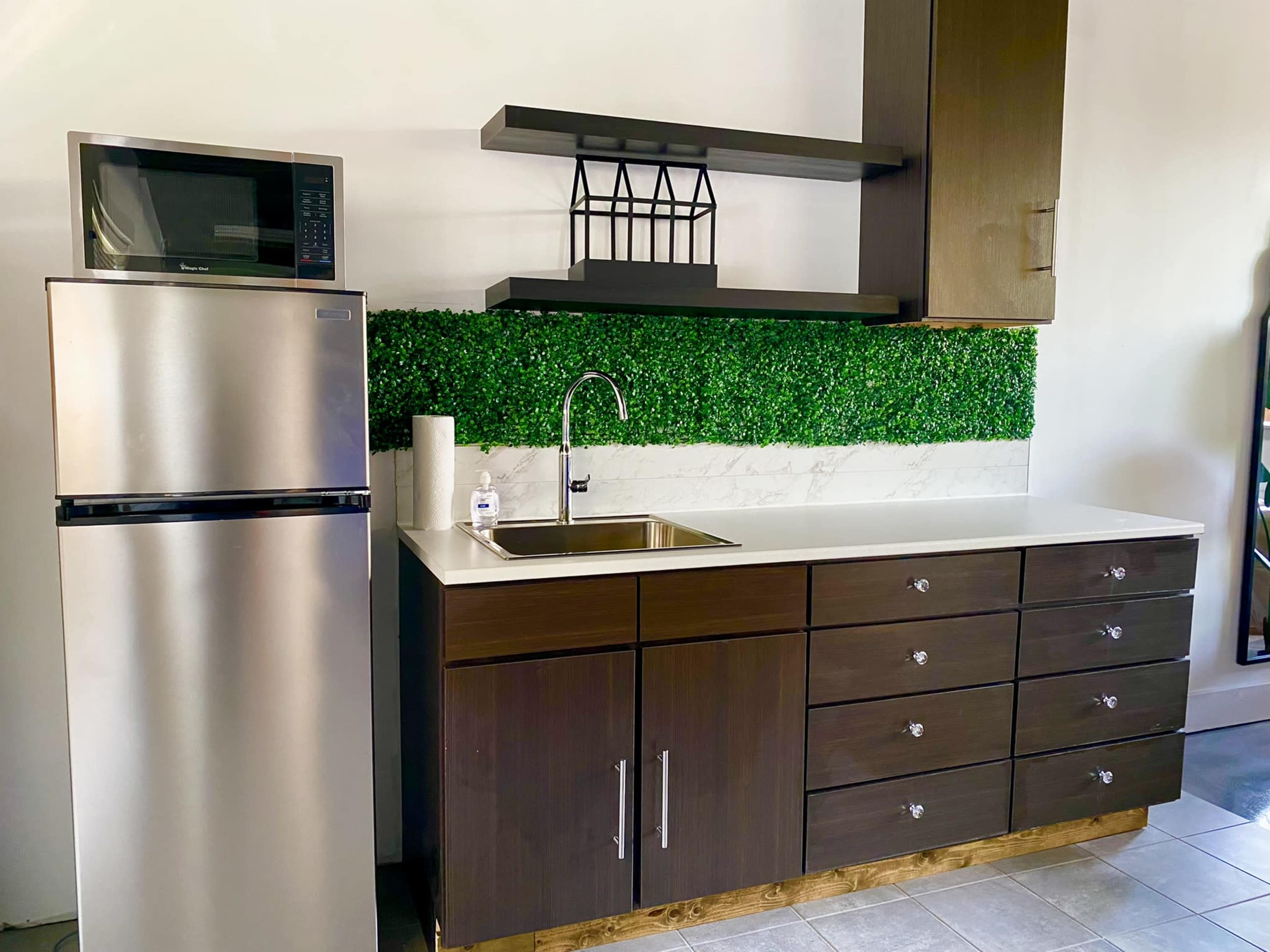 A modern kitchen with a stainless steel refrigerator, a sink with a faucet, and dark wood cabinets beneath a shelf featuring greenery.