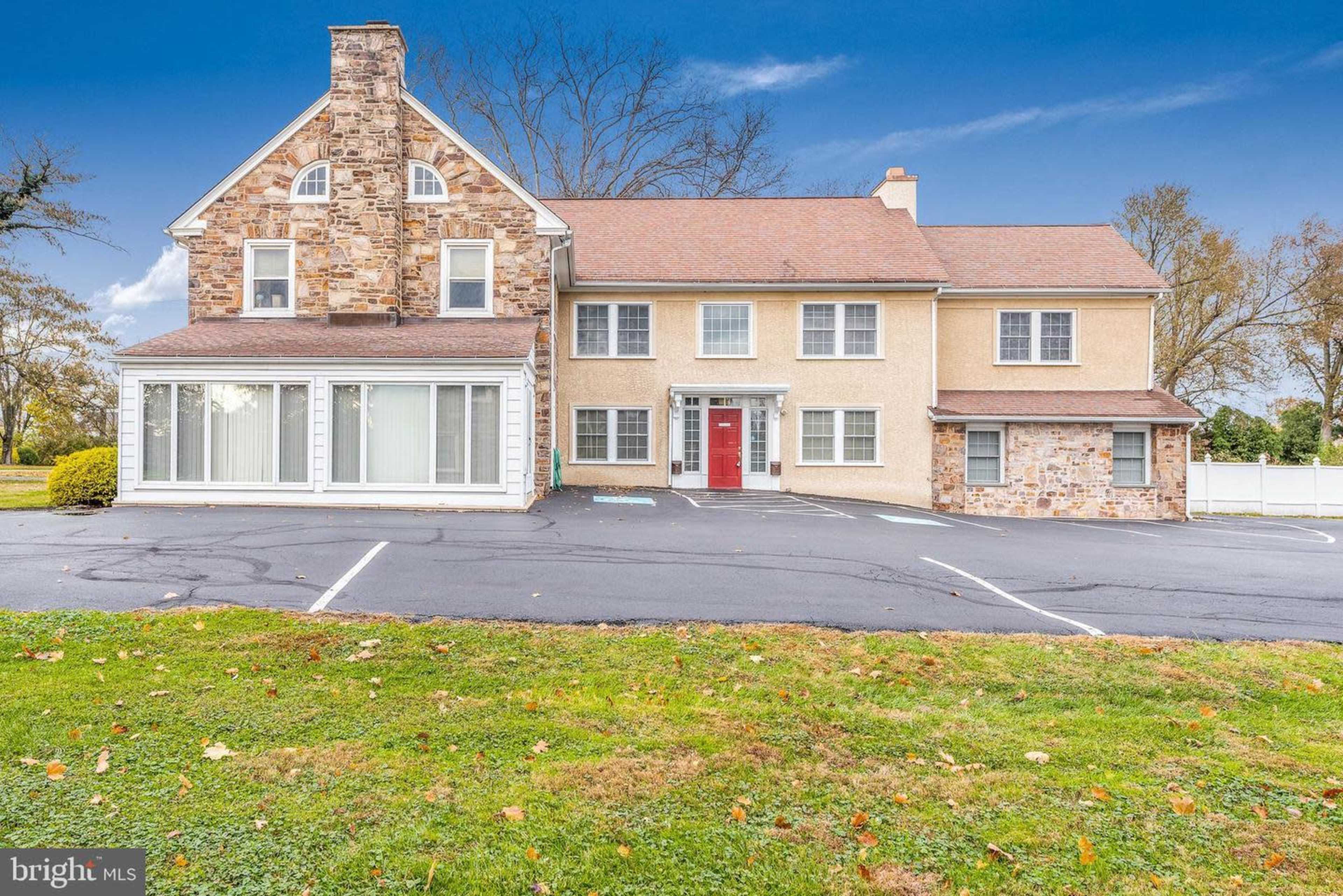 A large, two-story residential building with a combination of stone and stucco exteriors, featuring a red front door and a wraparound porch.