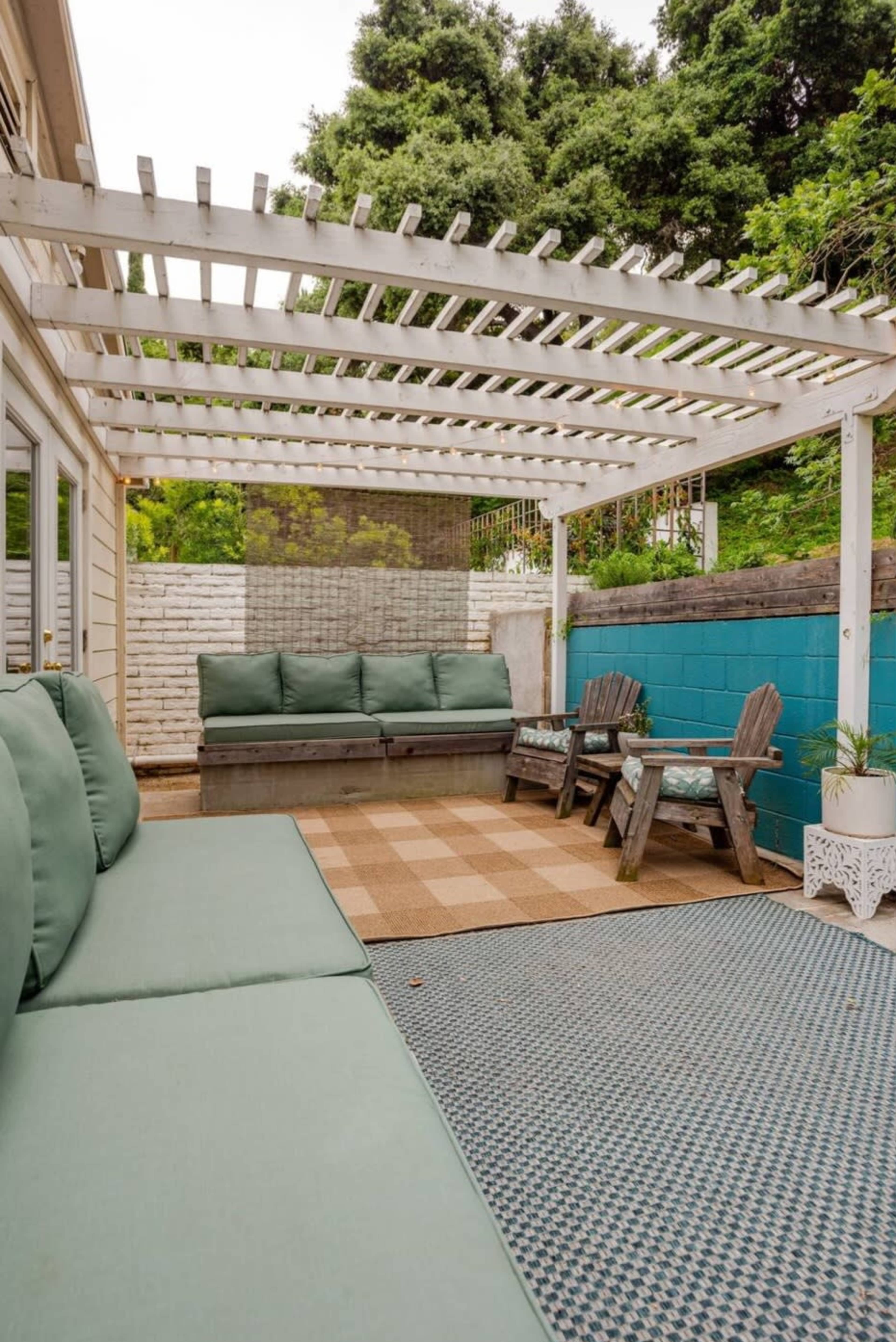A patio area features a slatted pergola, green seating cushions, a checkered rug, and wooden chairs against a blue and neutral wall.
