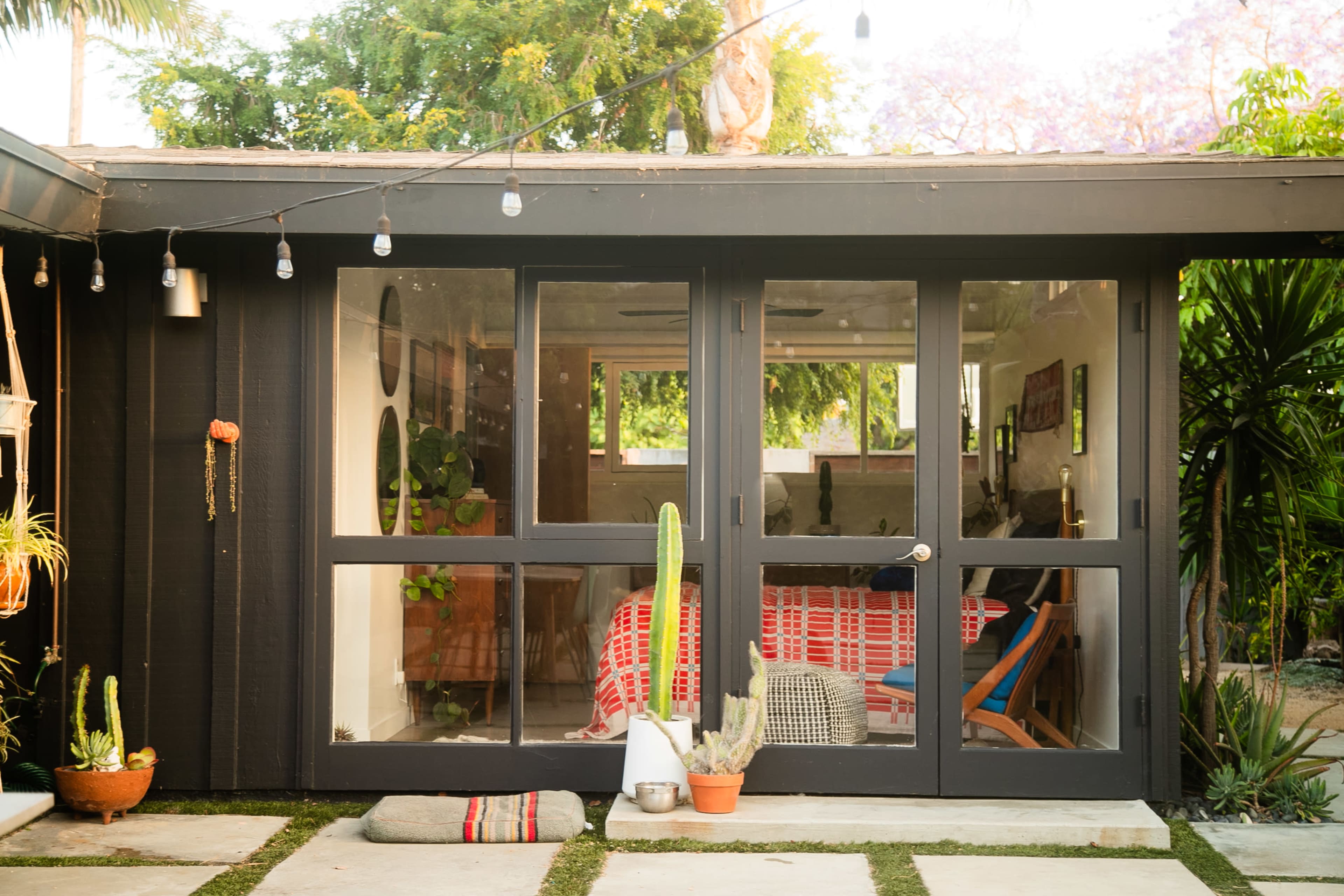 The image shows a modern, black wooden house with large glass doors leading into a brightly decorated interior, surrounded by potted plants and outdoor lighting.