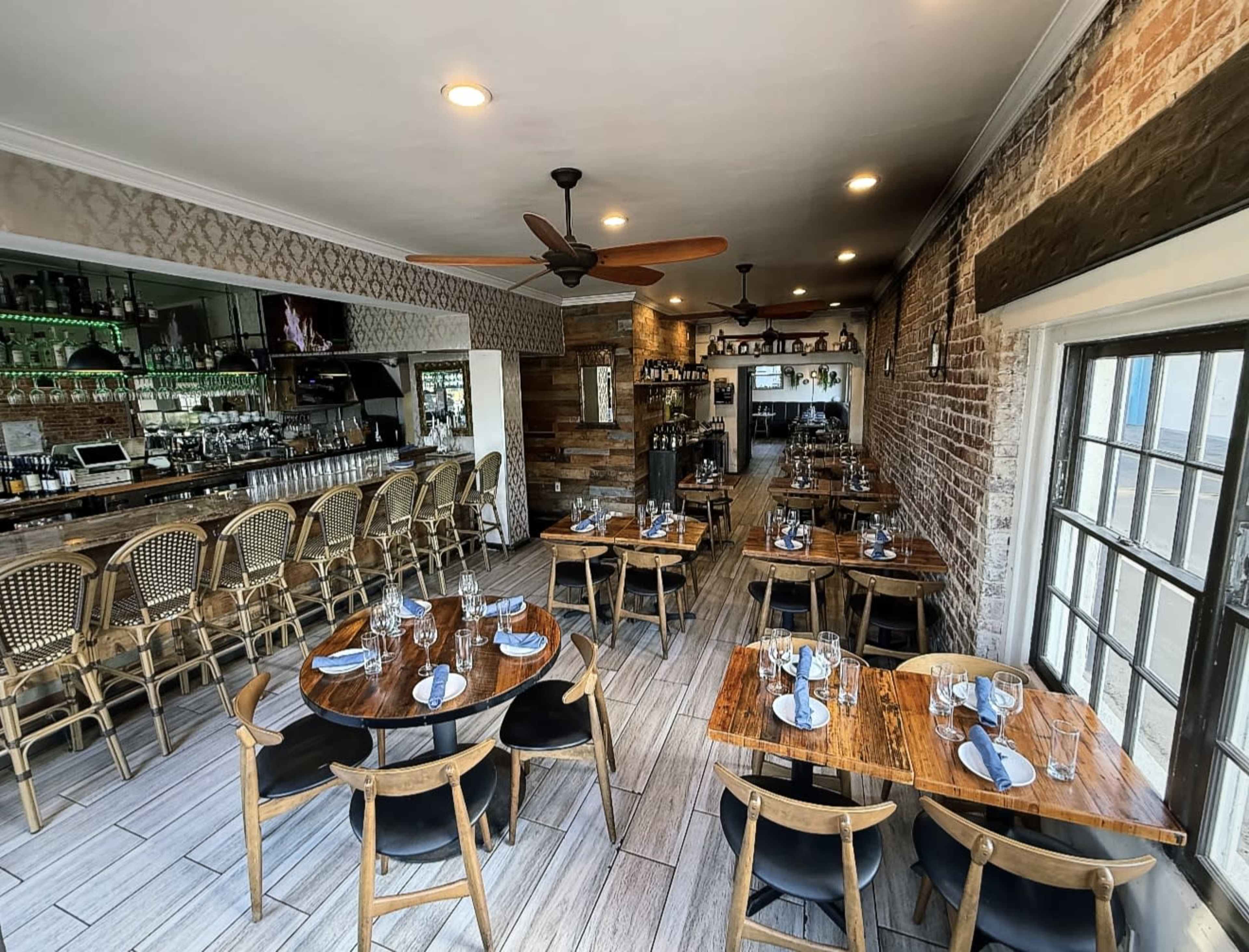 The image shows a restaurant interior featuring wooden tables set with blue napkins, surrounded by chairs, and a bar area with shelves of bottles in the background.