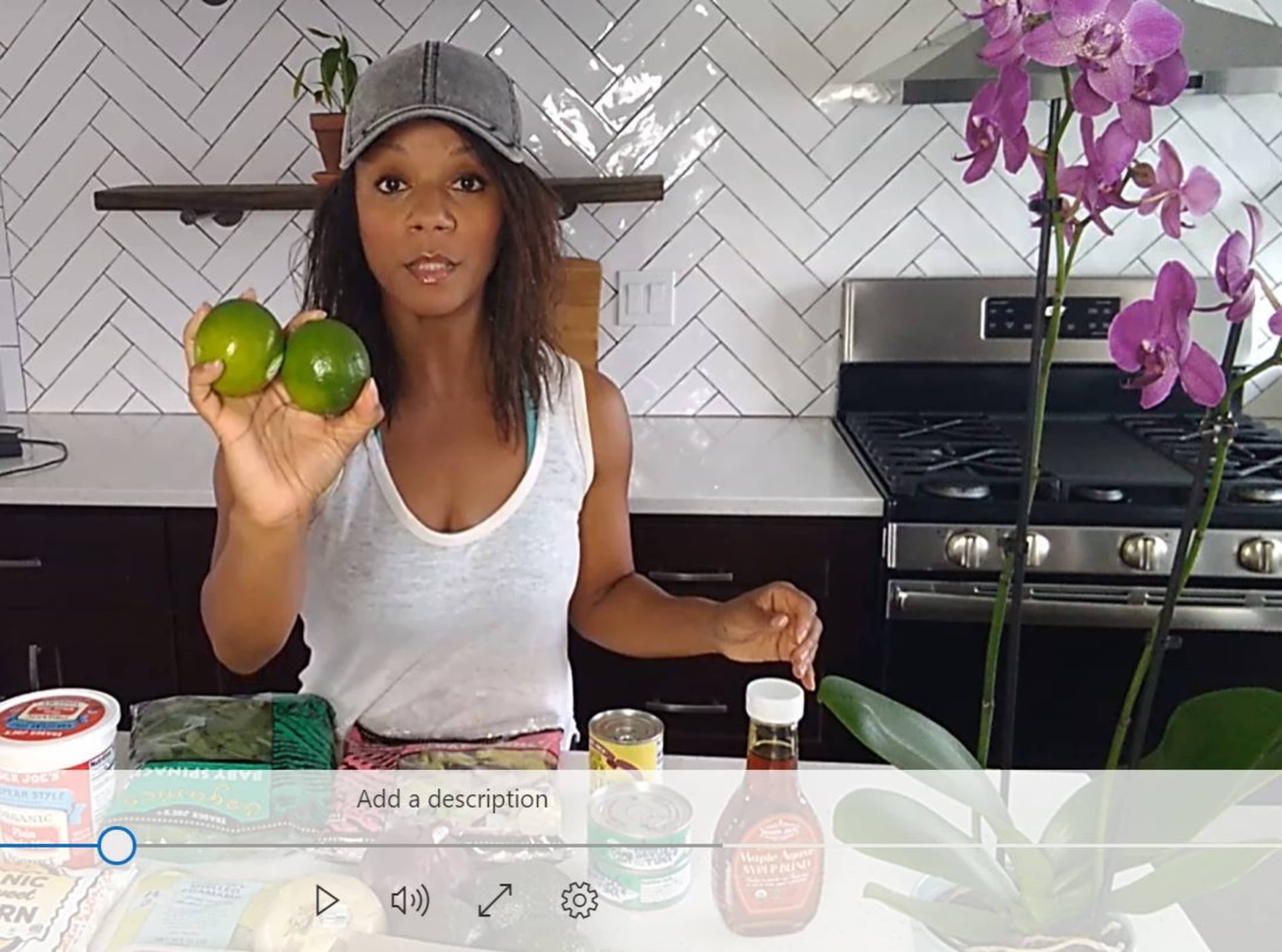 A person is holding two limes in a kitchen while surrounded by various food items and a potted orchid.