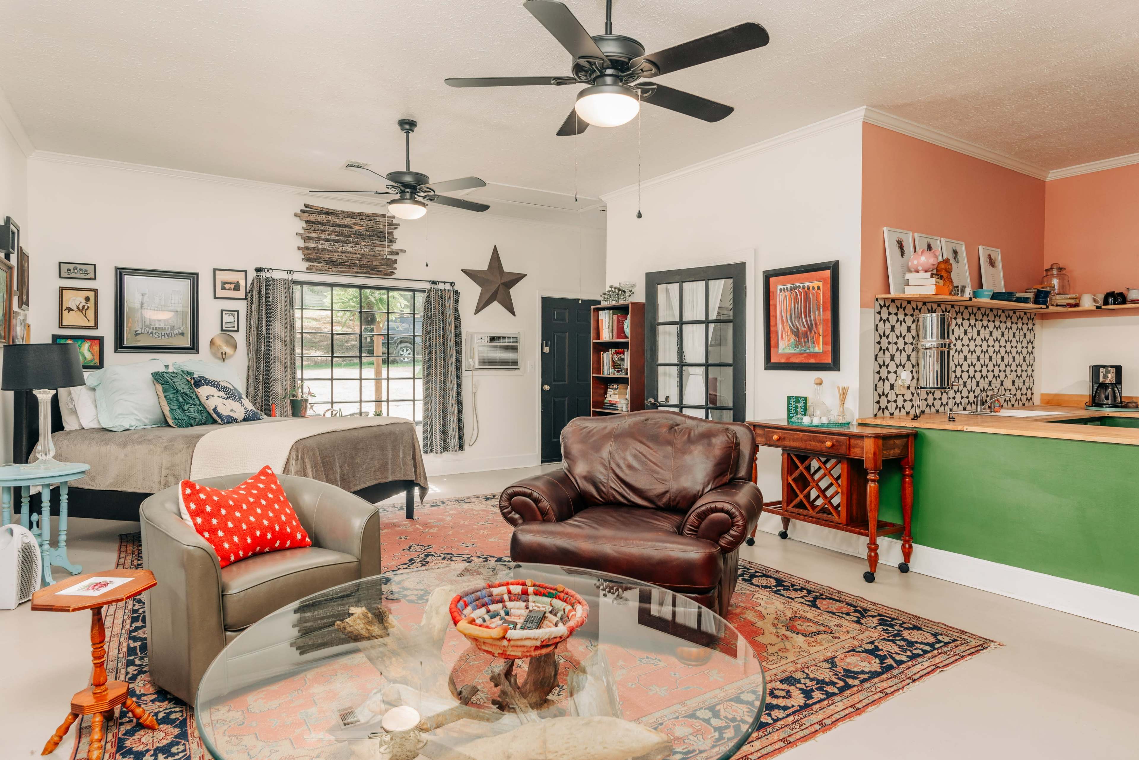 The image shows a cozy living space with a bed, a seating area, and a small kitchen featuring green cabinetry and a decorative wall.