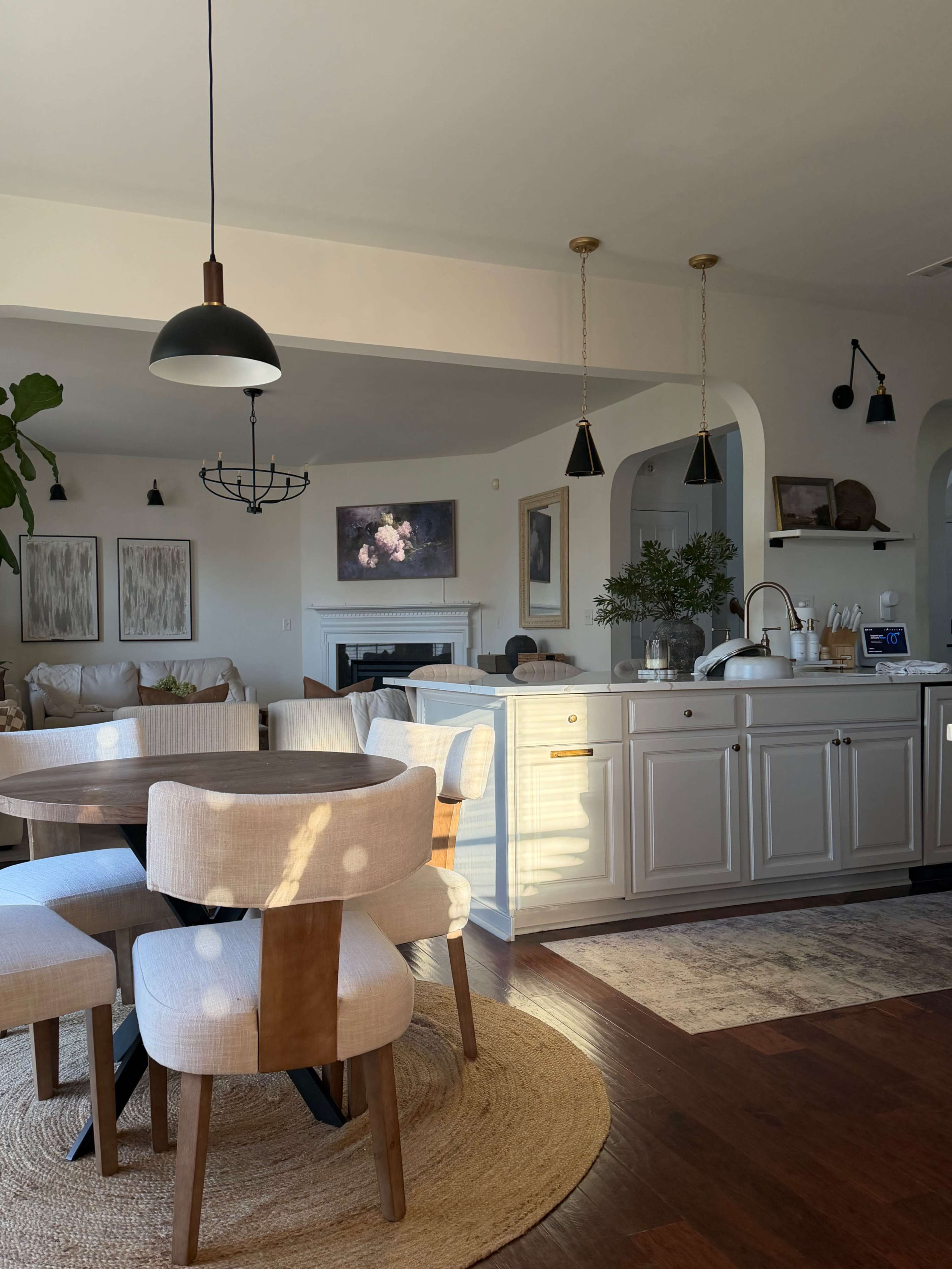 The image shows a bright, open-concept living area featuring a circular dining table surrounded by upholstered chairs, a modern kitchen with light cabinetry, and decorative wall art.