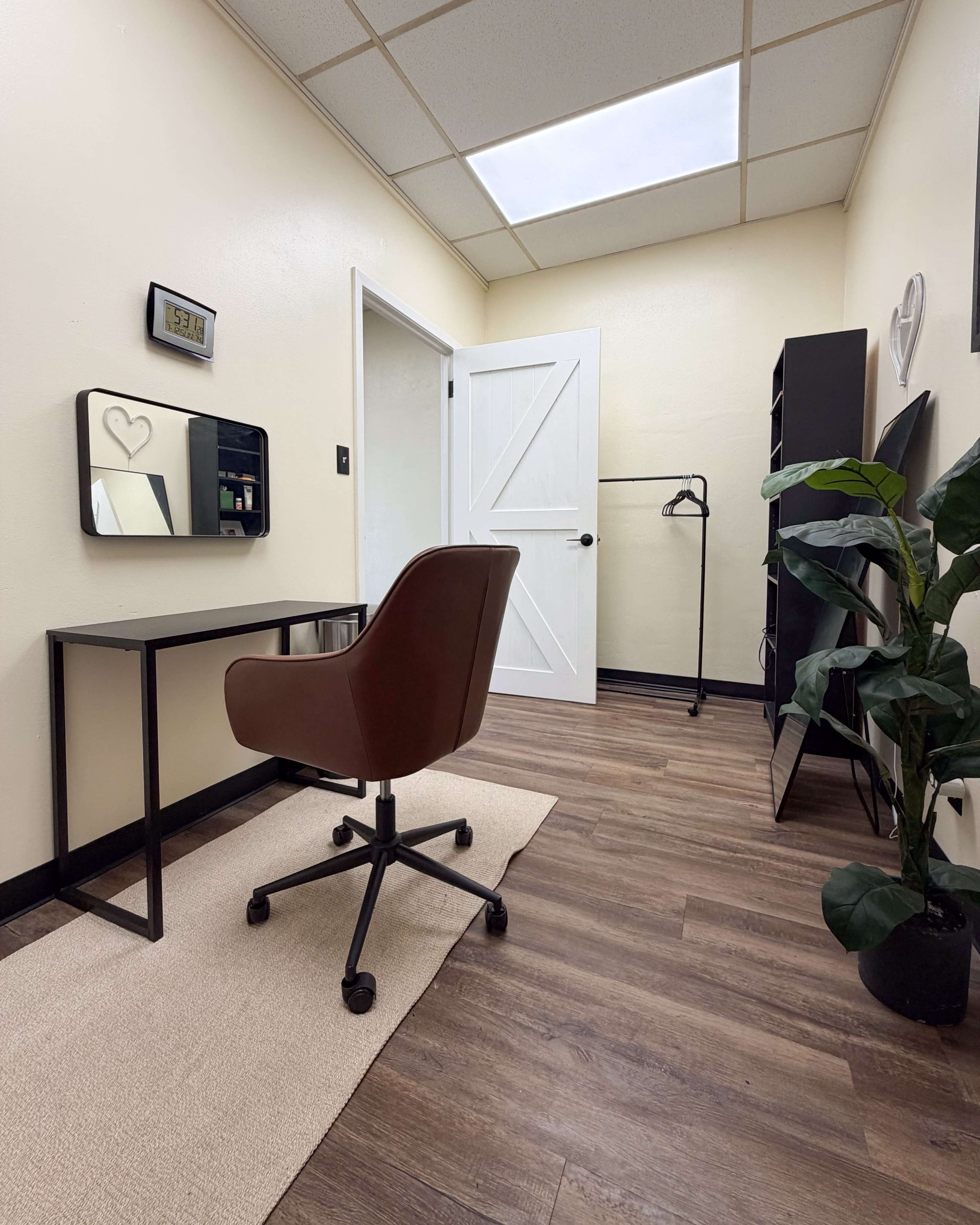 The image shows a small, well-lit room with a desk, chair, mirror, plant, and a door leading to another area.