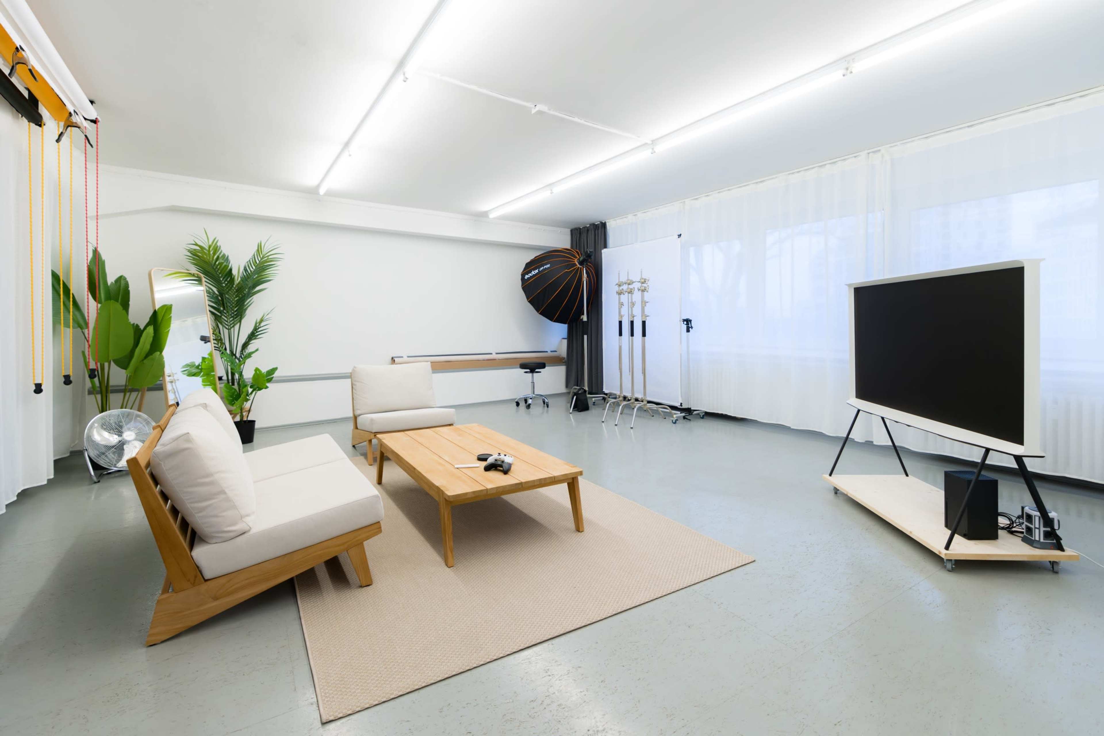 The image shows a bright, minimalist living space featuring a wooden coffee table, two light-colored sofas, and a large television against a plain wall, with plants and studio equipment in the background.
