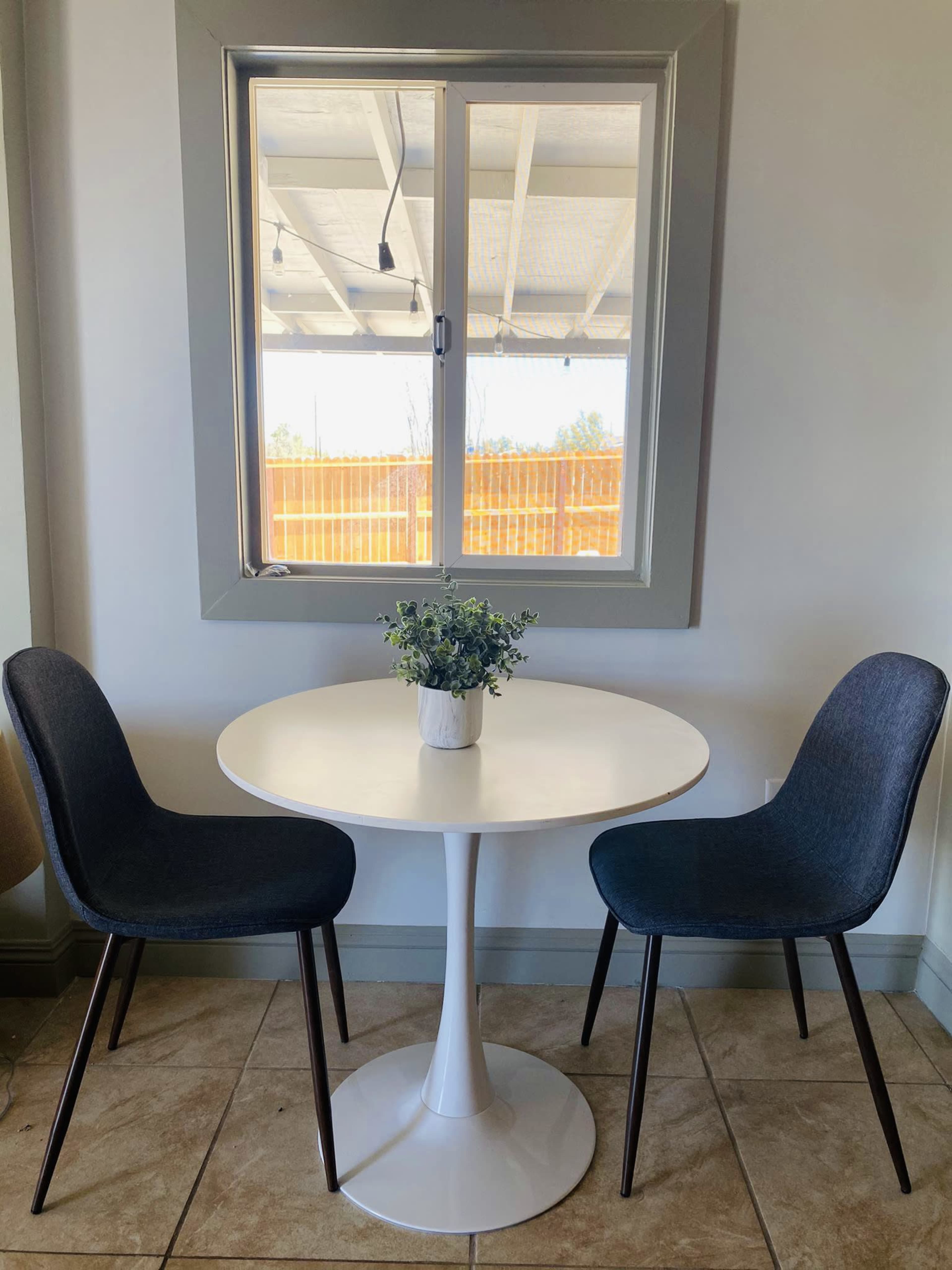A round white table with a small vase of flowers sits between two dark chairs under a window.