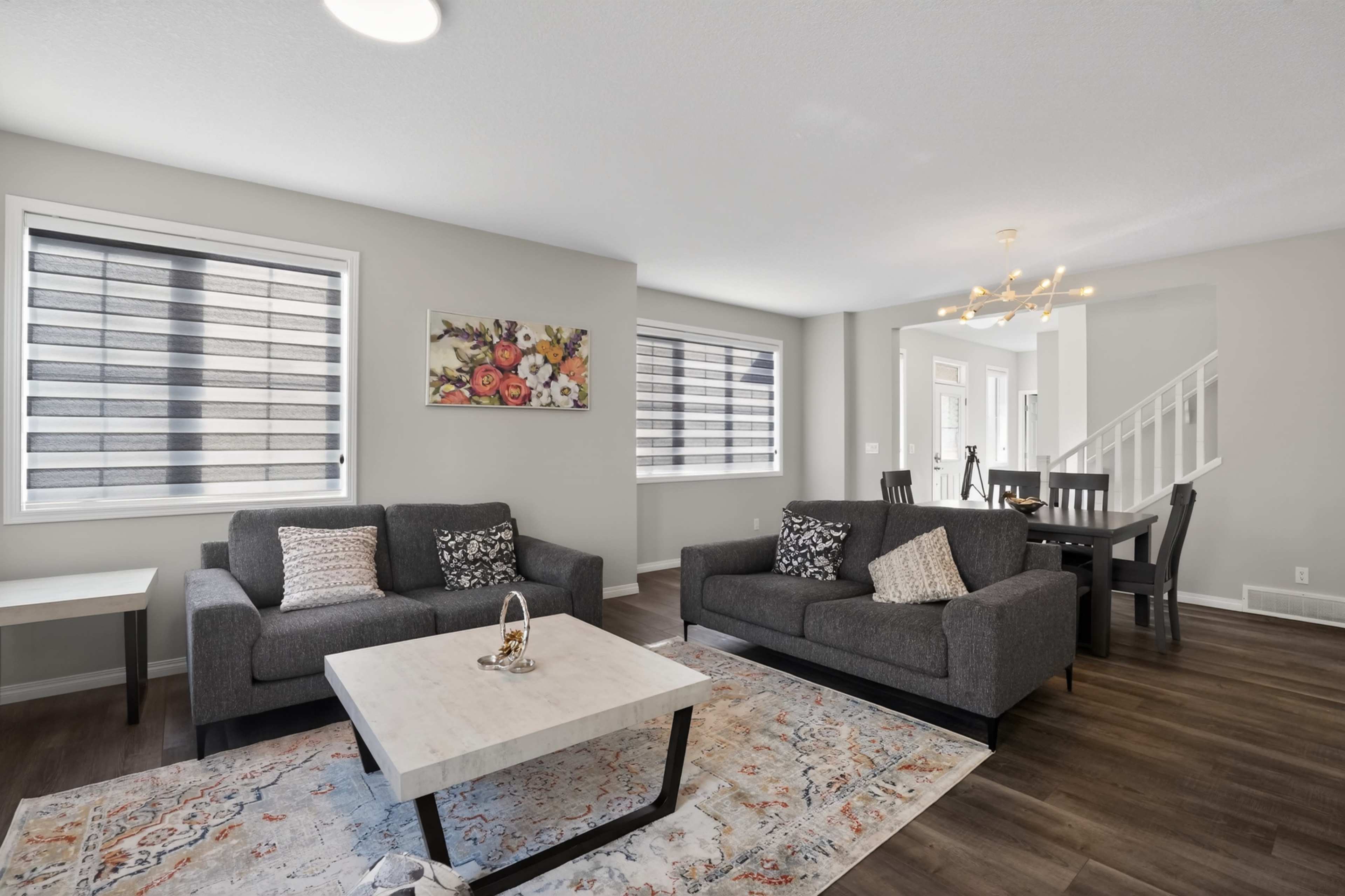 The image shows a modern living room with gray sofas, a marble coffee table, a floral wall art piece, and large windows dressed with blinds, adjacent to a dining area.