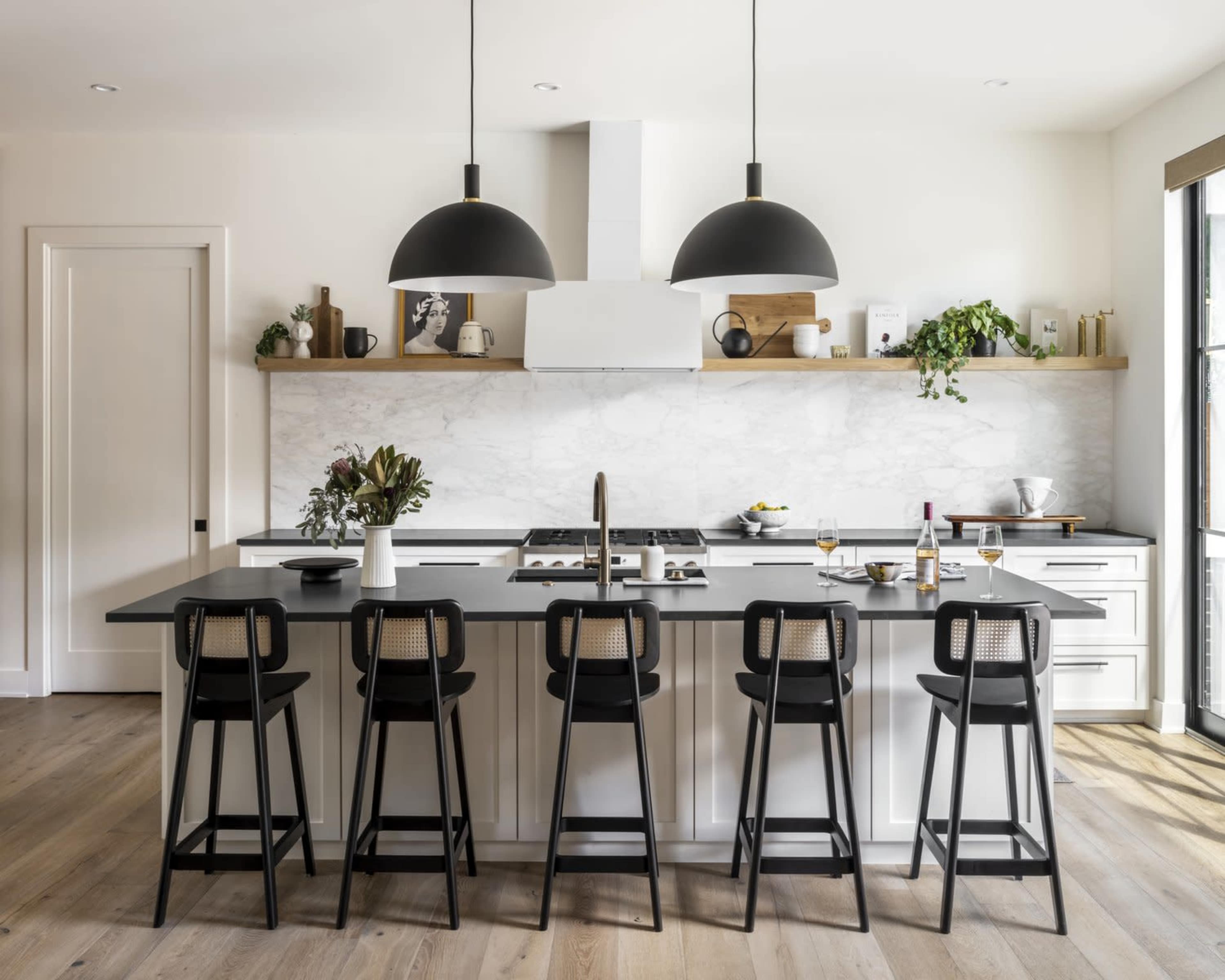 A modern kitchen features a large island with six black bar stools, two pendant lights overhead, and a marble backsplash.
