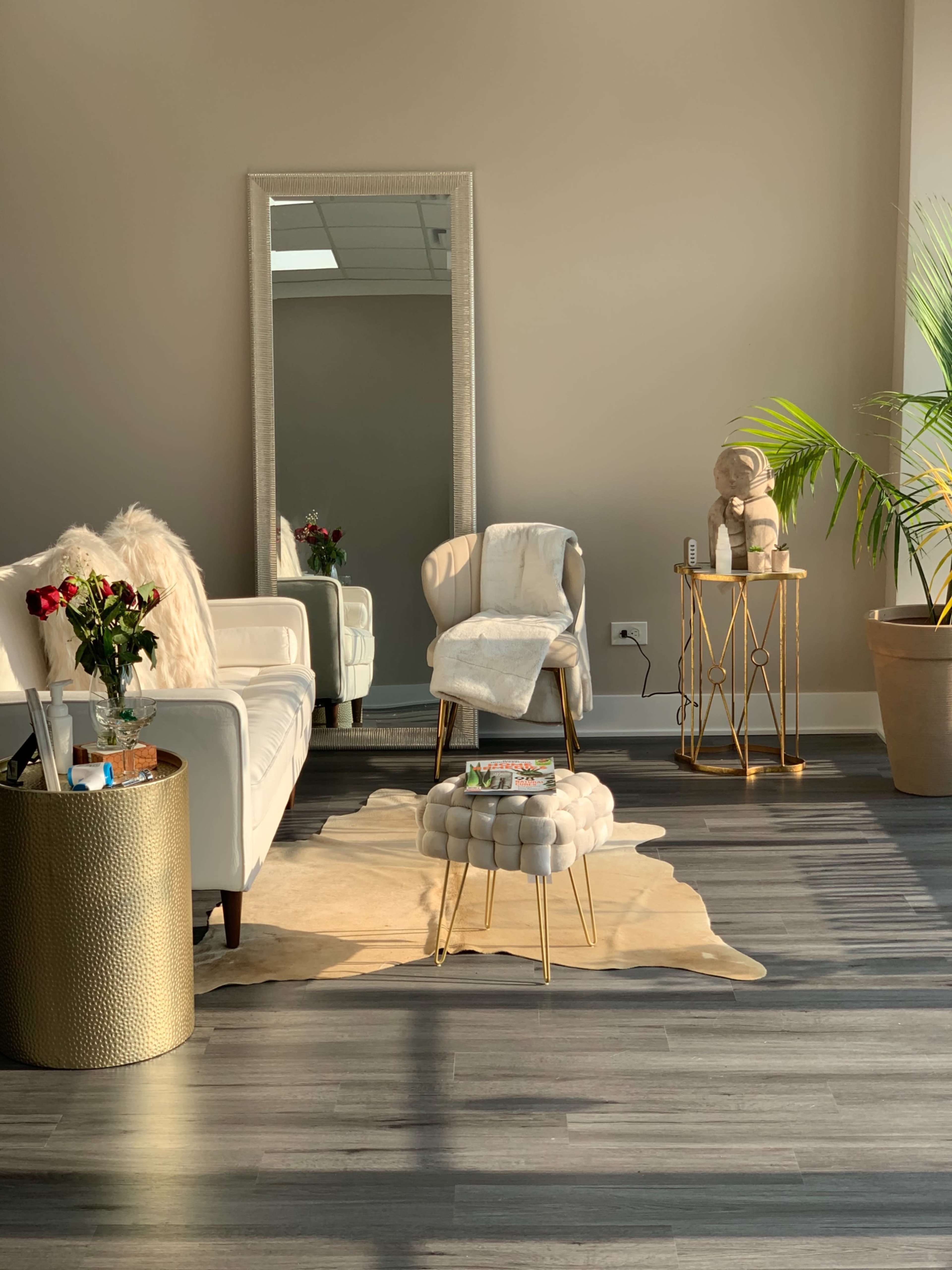 A modern waiting area features a white sofa, a gold-accented side table, a cowhide rug, and a large mirror reflecting natural light.