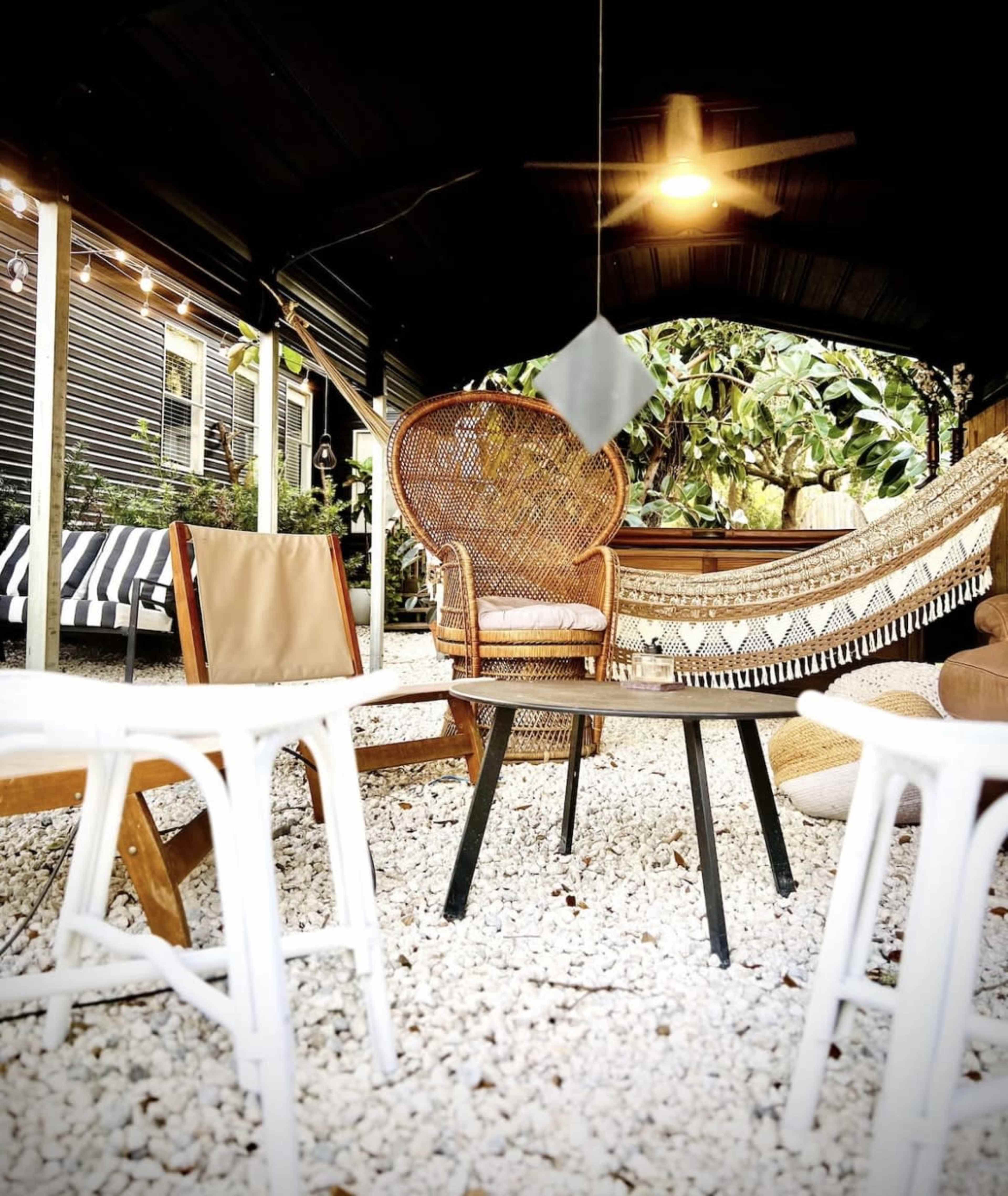 A patio area with a hammock, a wicker chair, and several small tables surrounded by gravel and lush greenery.