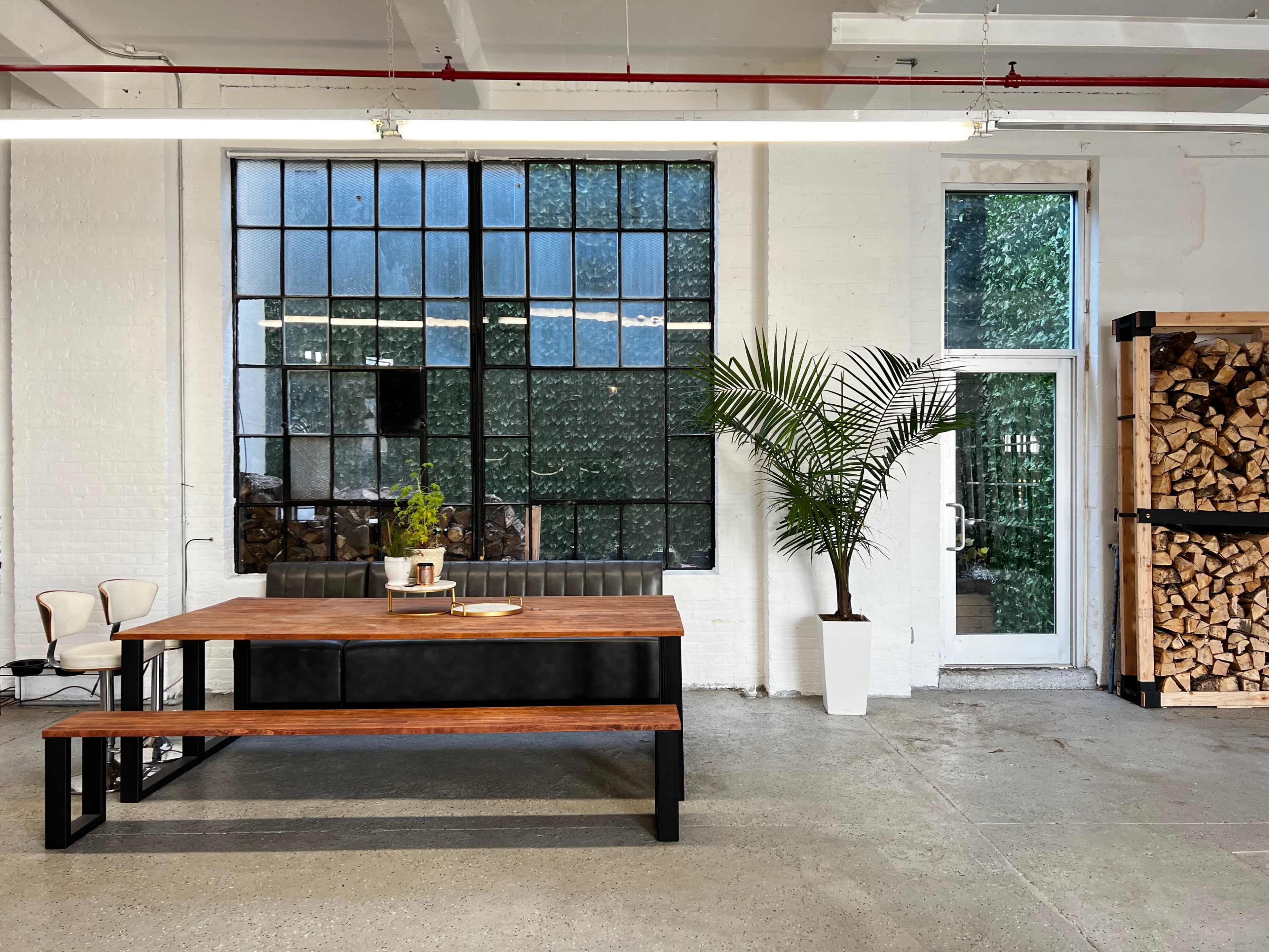 A spacious room with a large wooden table and benches, a potted palm plant, a stacked wood pile, and tall windows allowing natural light to enter.