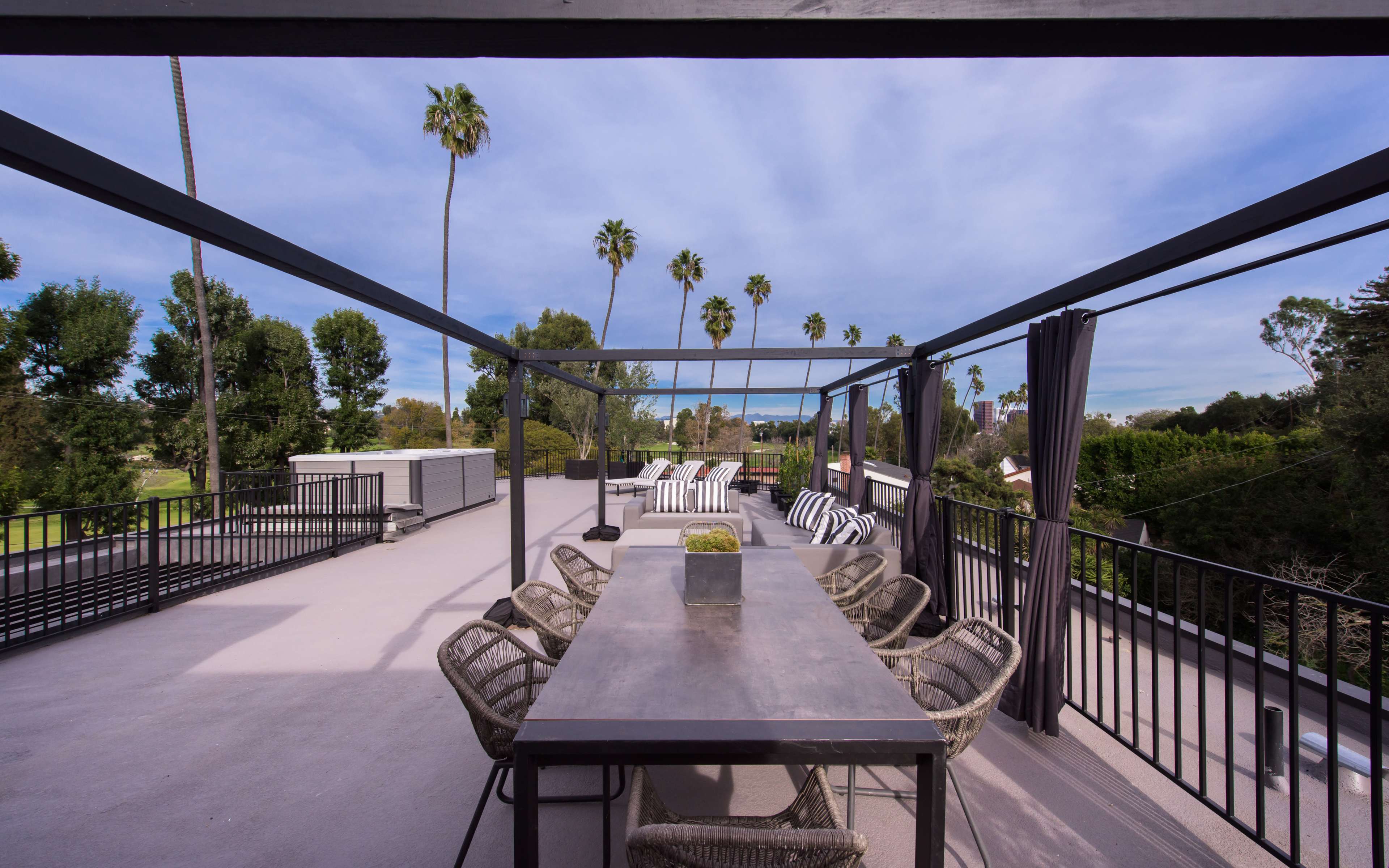 A spacious rooftop terrace features a long dining table surrounded by chairs, with palm trees visible in the background under a partly cloudy sky.