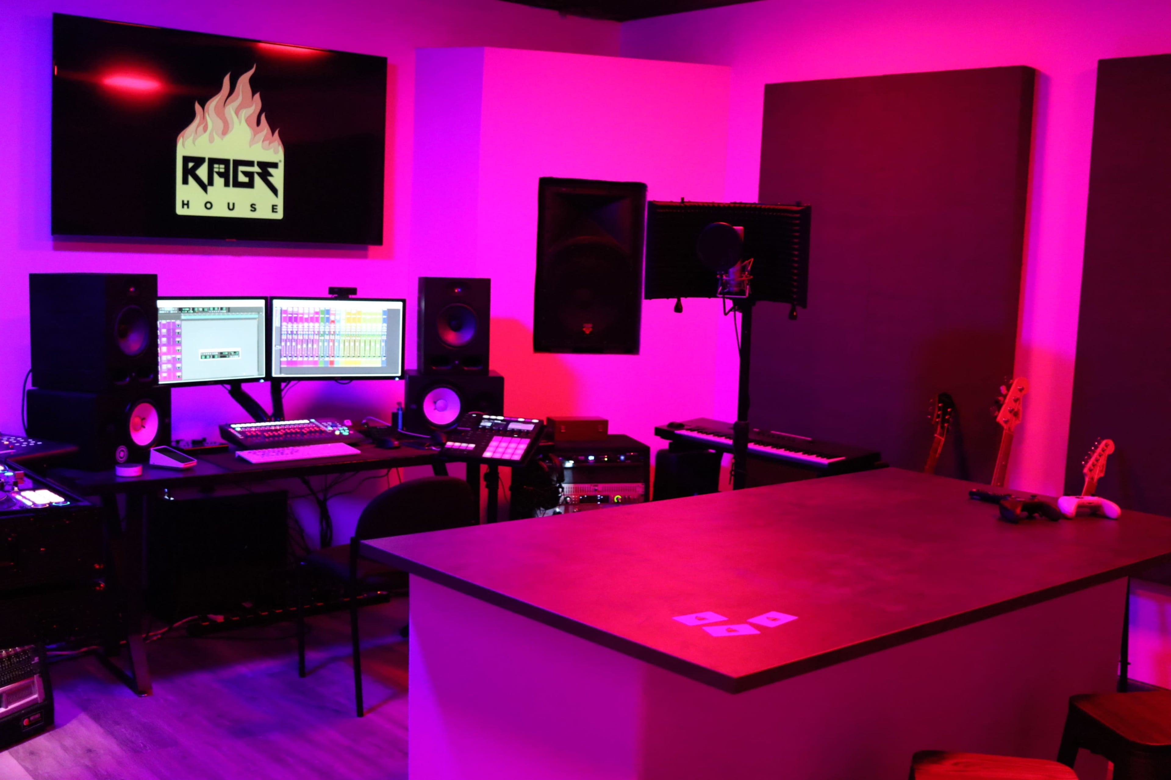 A music production studio with a desk, two computer monitors, audio equipment, speakers, and an illuminated wall sign labeled "RAGE HOUSE."