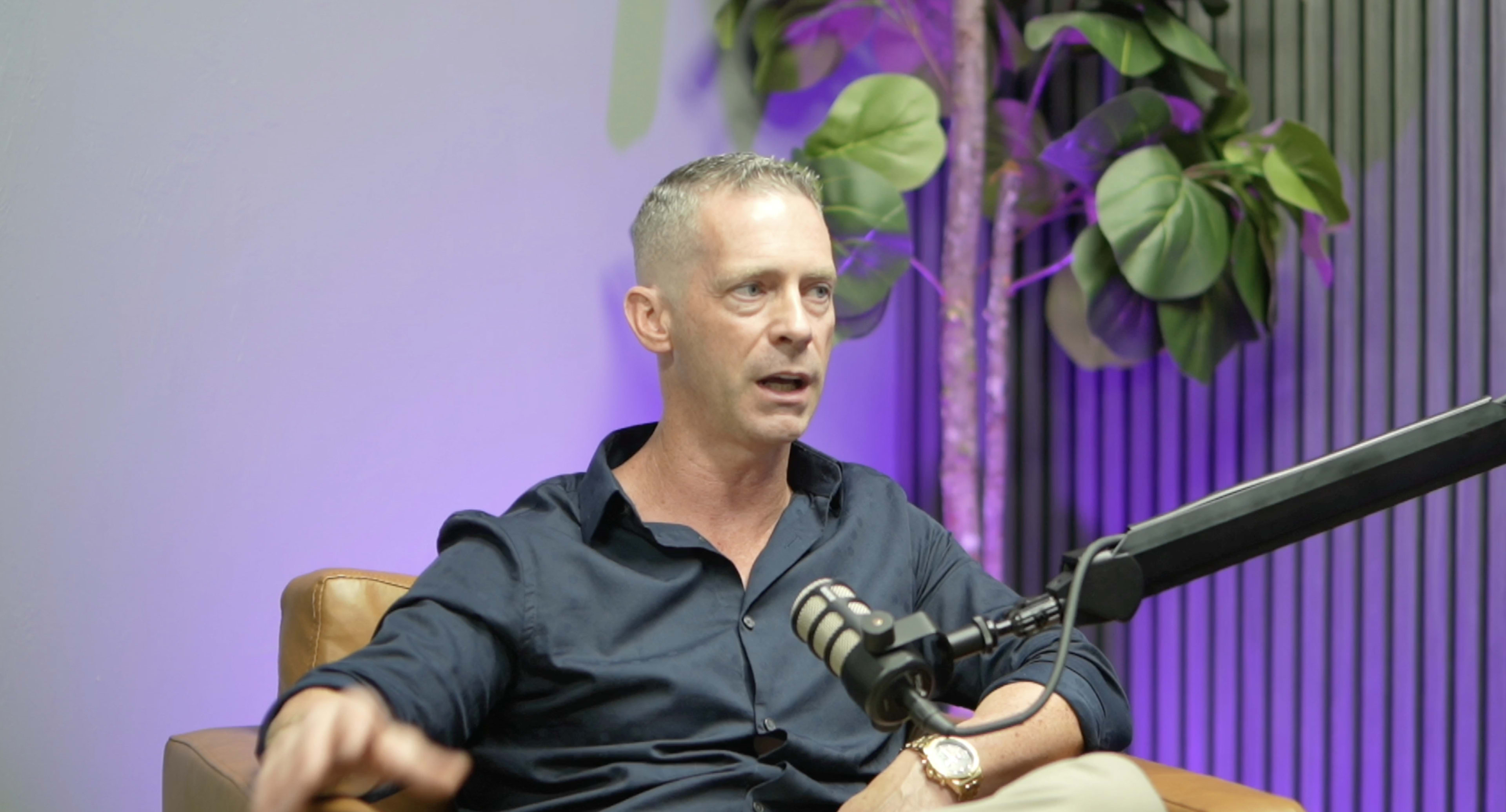 A man in a dark shirt is speaking while seated on a brown couch, with a microphone in front of him and plants in the background.