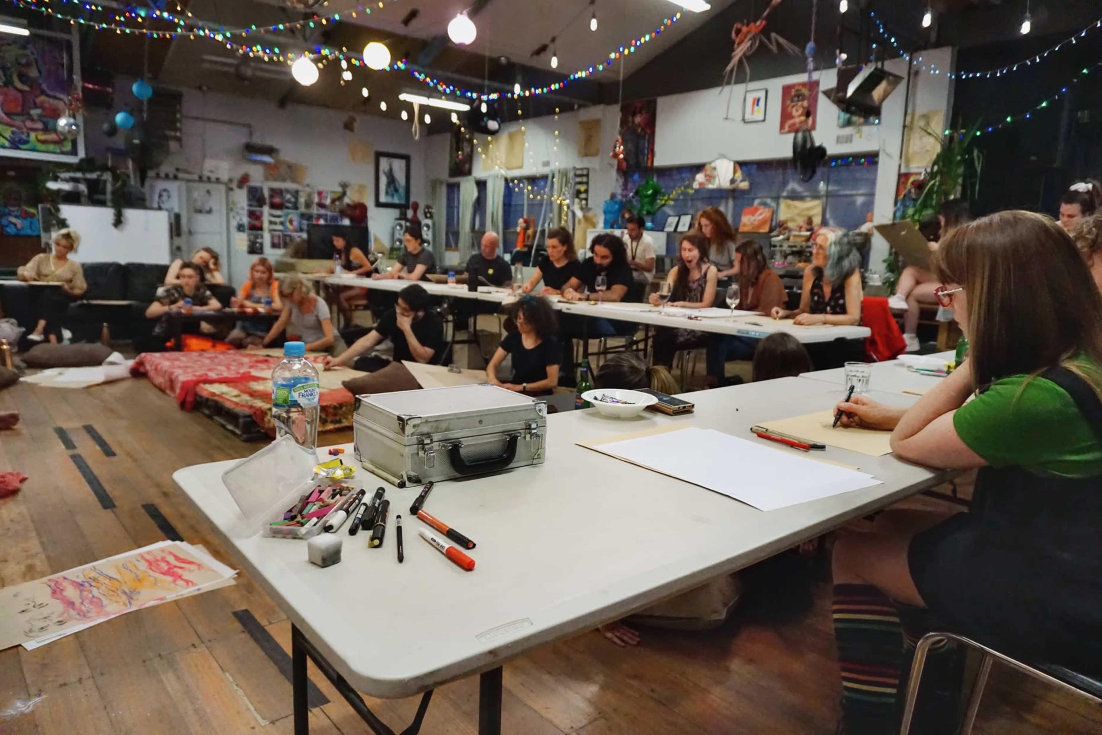 A group of people sits at tables in a brightly lit art studio, engaged in drawing and painting activities.