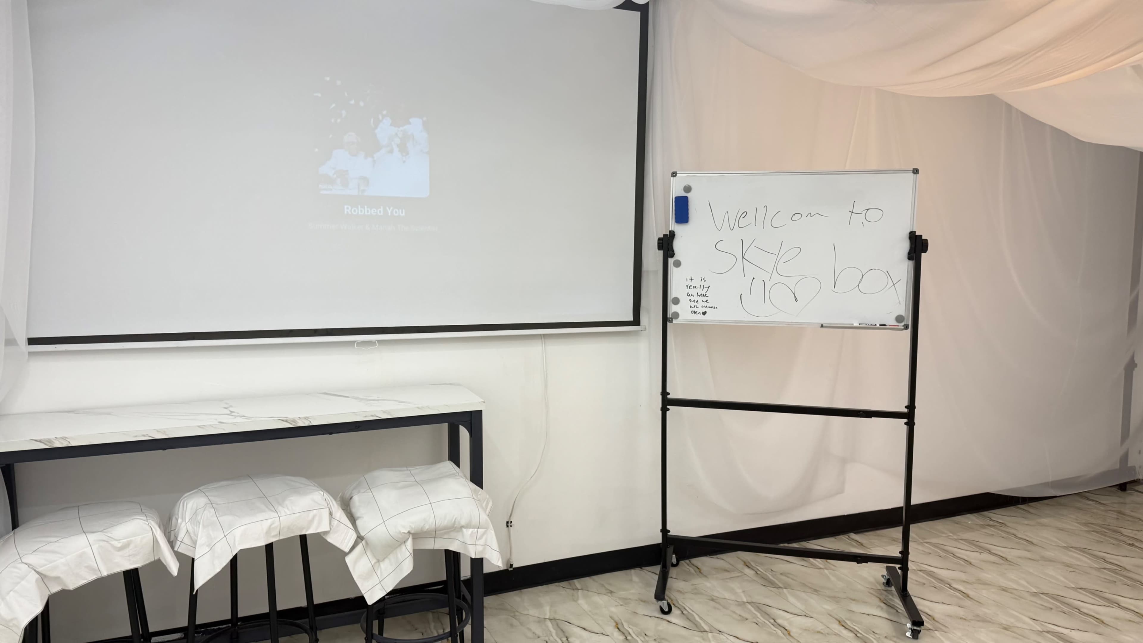 A projector screen displays a slide, while a whiteboard beside it has a handwritten message welcoming people to a sky box.
