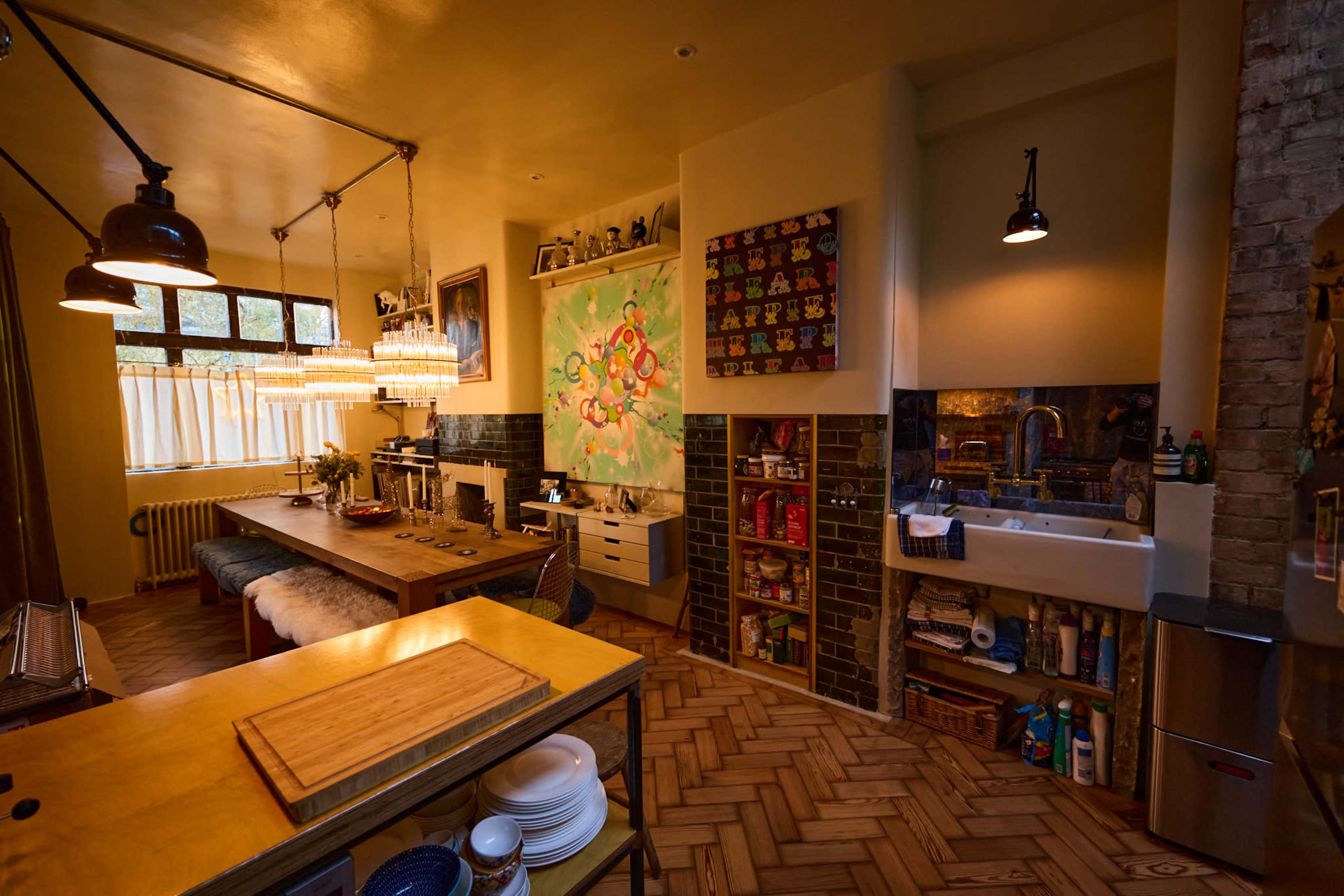 The image shows a modern kitchen and dining area with a large wooden table, colorful artwork on the walls, and shelves filled with various kitchen items.