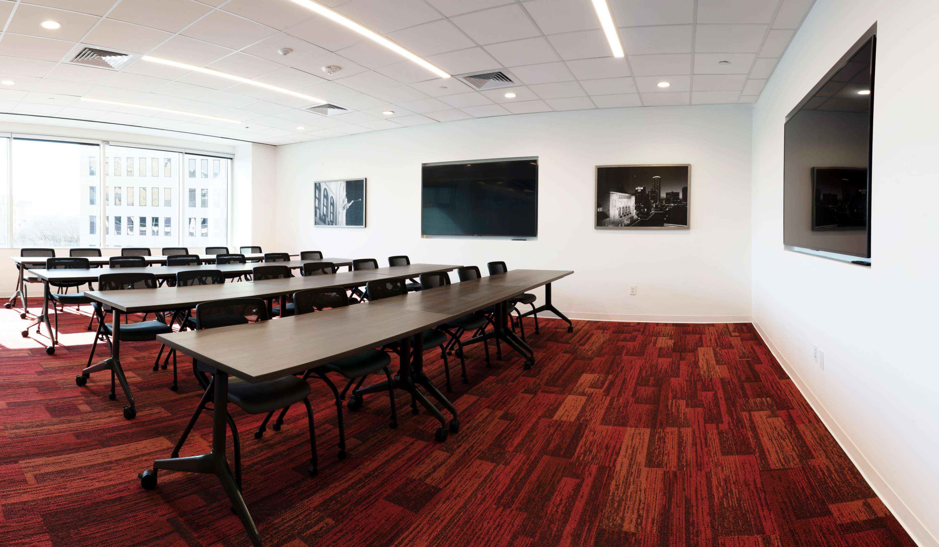 A conference room features multiple long tables arranged for seating, with two large screens mounted on the walls and photographs displayed.