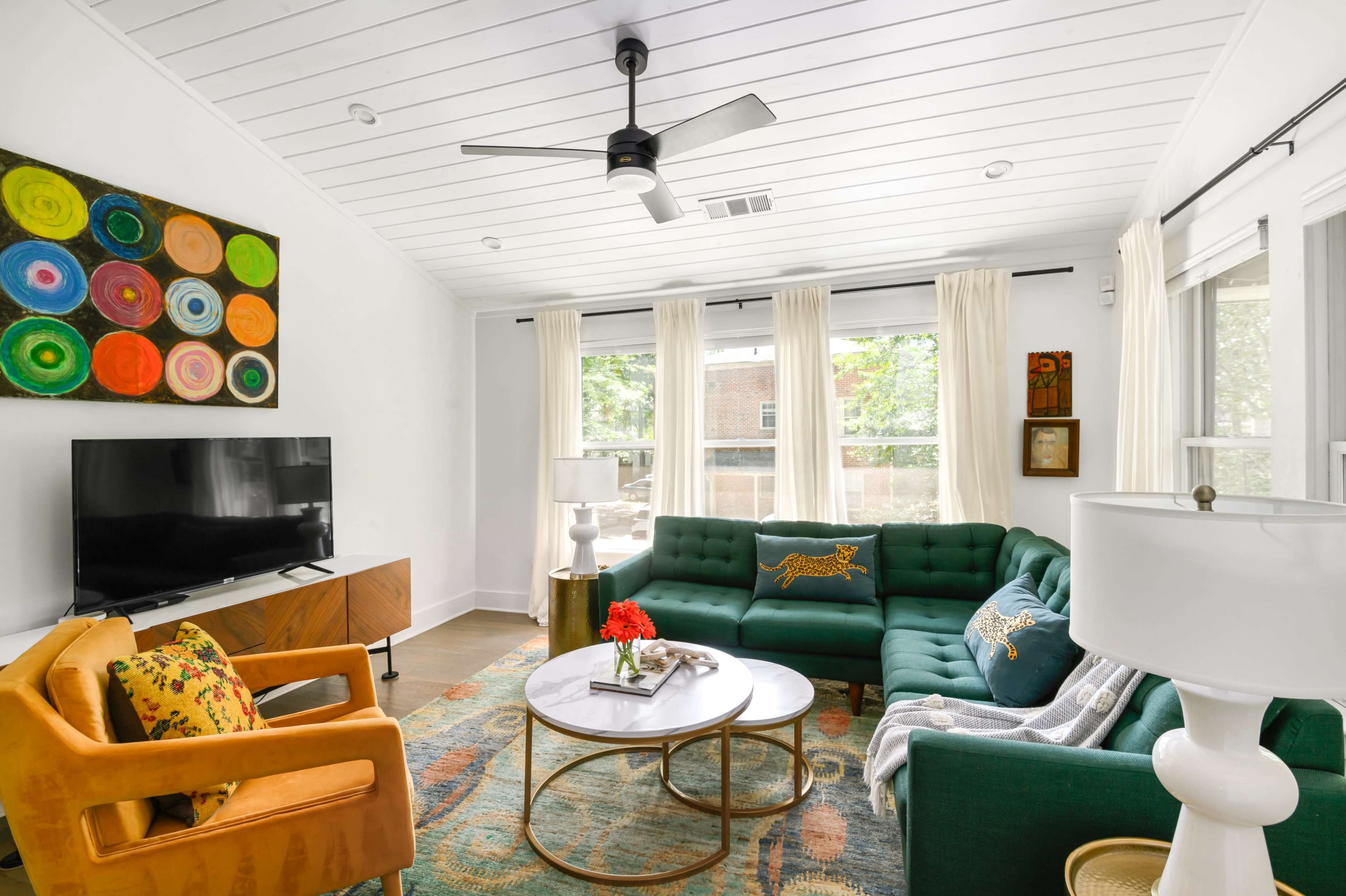 The image shows a modern living room featuring a green sectional sofa, a wooden TV stand with a television, a round coffee table, and colorful artwork on the wall, with large windows allowing natural light to enter.