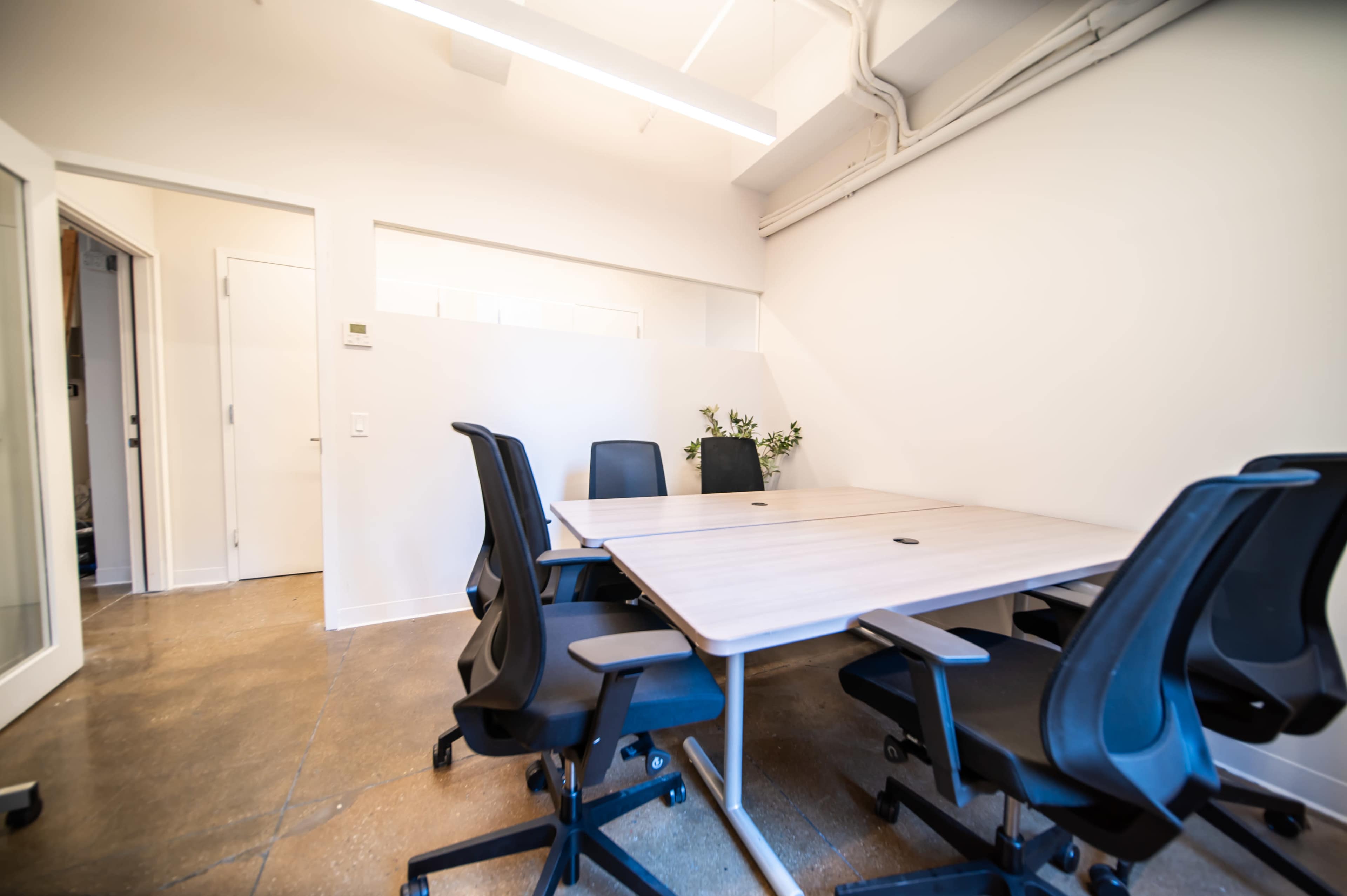 A simple meeting room features a large wooden table surrounded by black ergonomic chairs, with a door leading to another space in the background.
