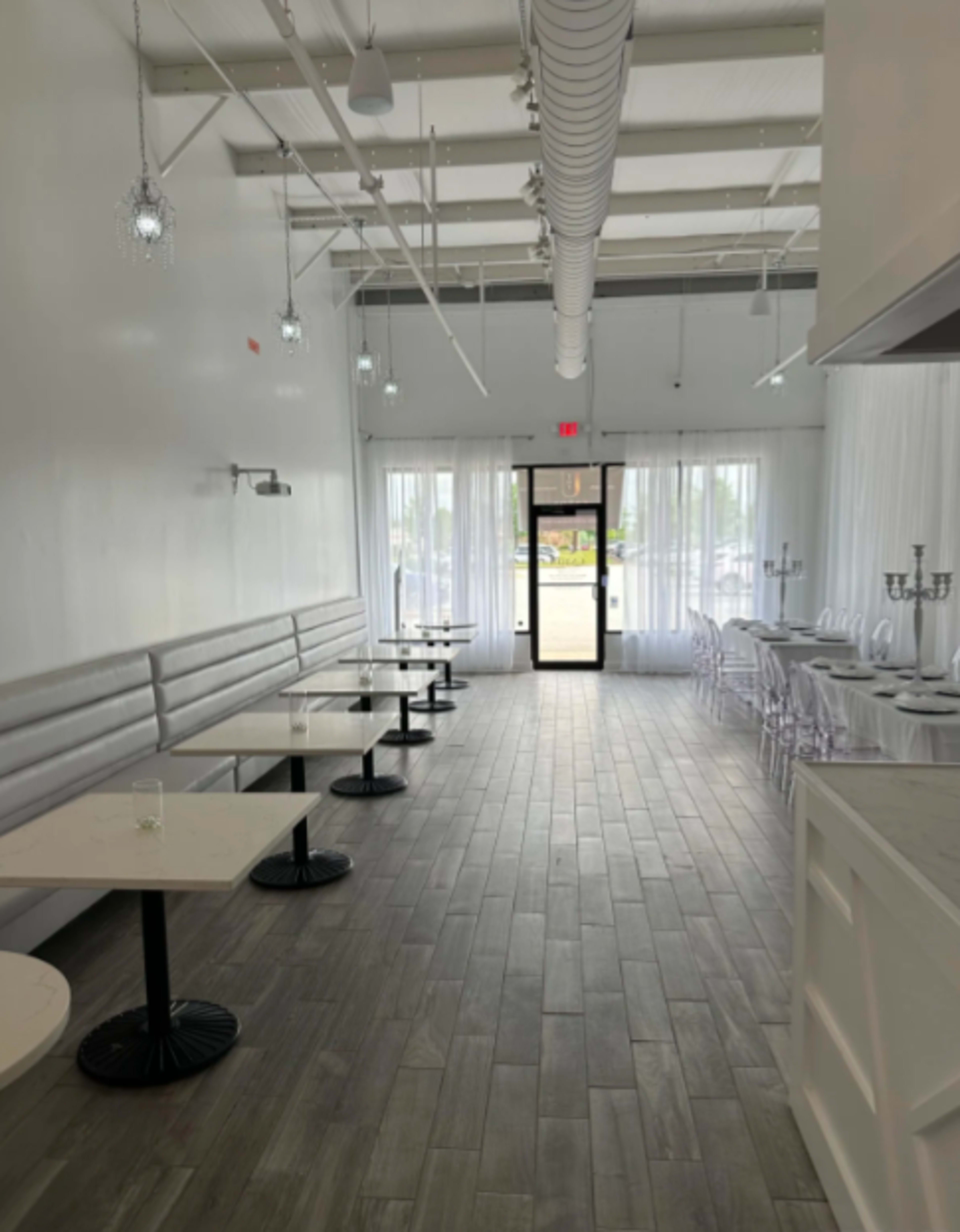 The image shows a modern, minimalist restaurant interior with white walls, wooden flooring, and several round tables along one side, leading to a glass door at the far end.