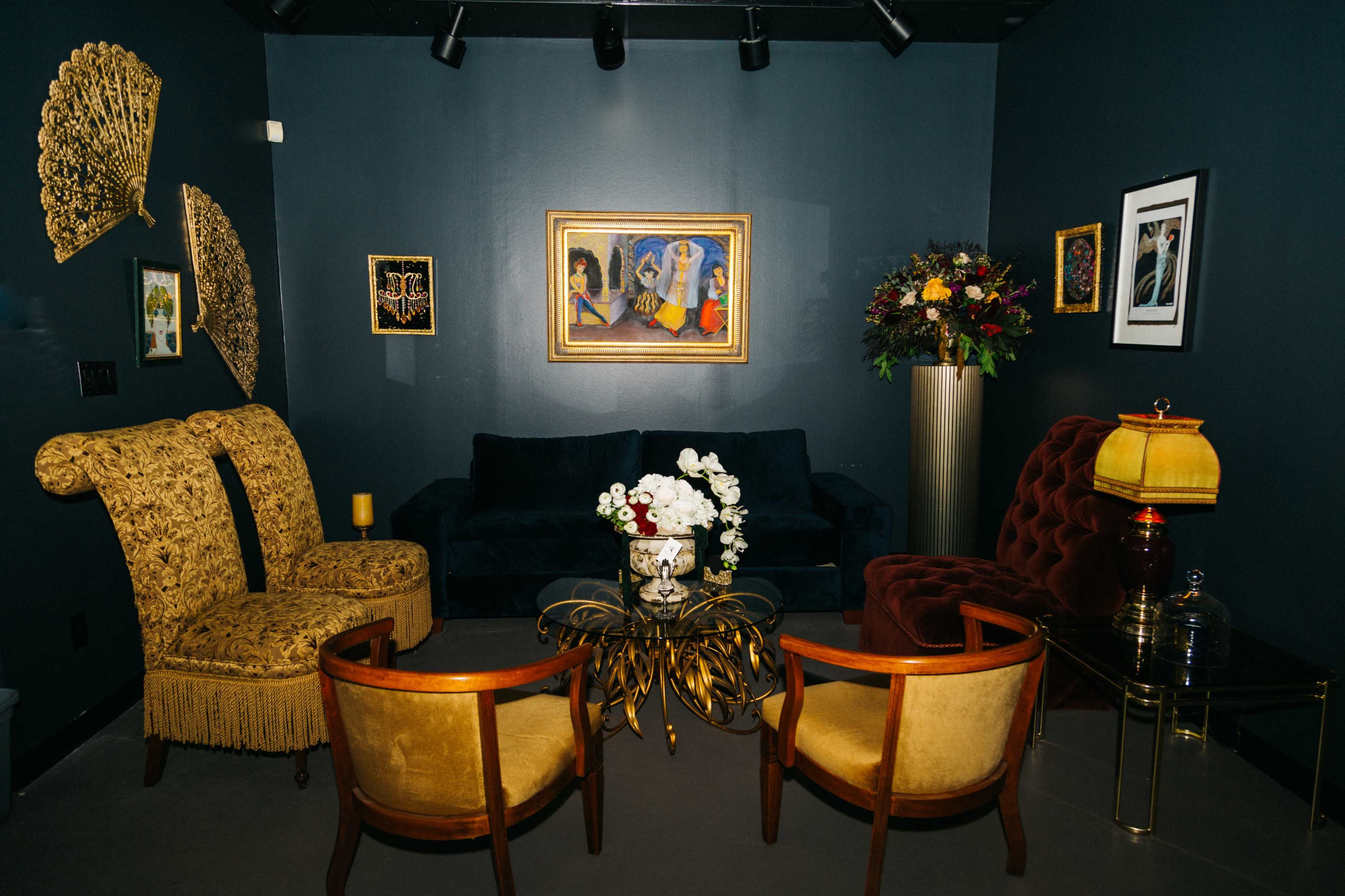 A stylish lounge area featuring two ornate chairs, a velvet sofa, a gold-accented coffee table, and decorative wall art.