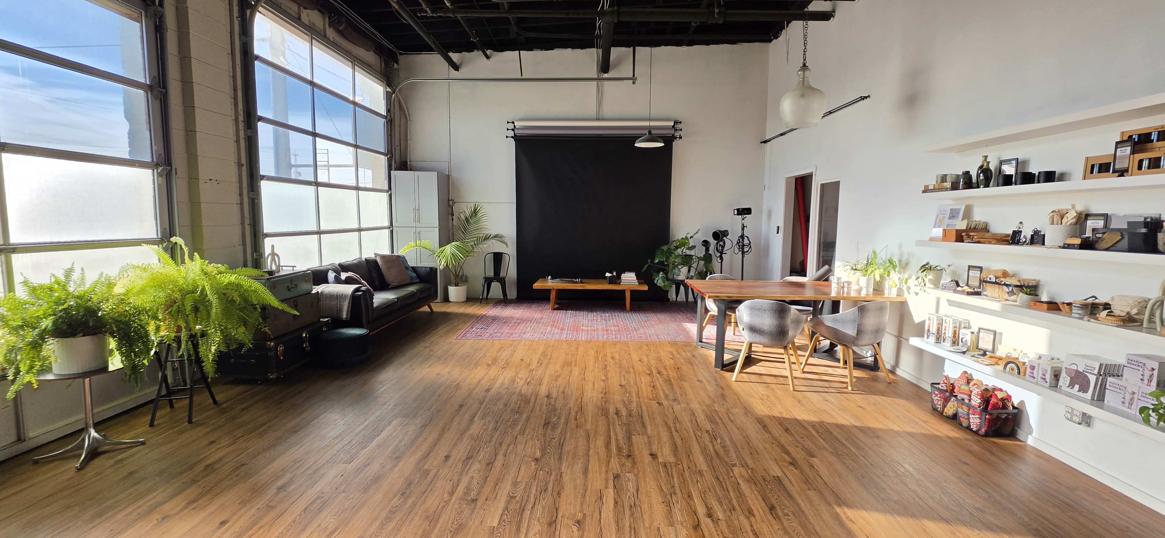 The image shows a spacious, well-lit studio with large windows, a black backdrop for photography, a wooden table, and plants decorating the area.