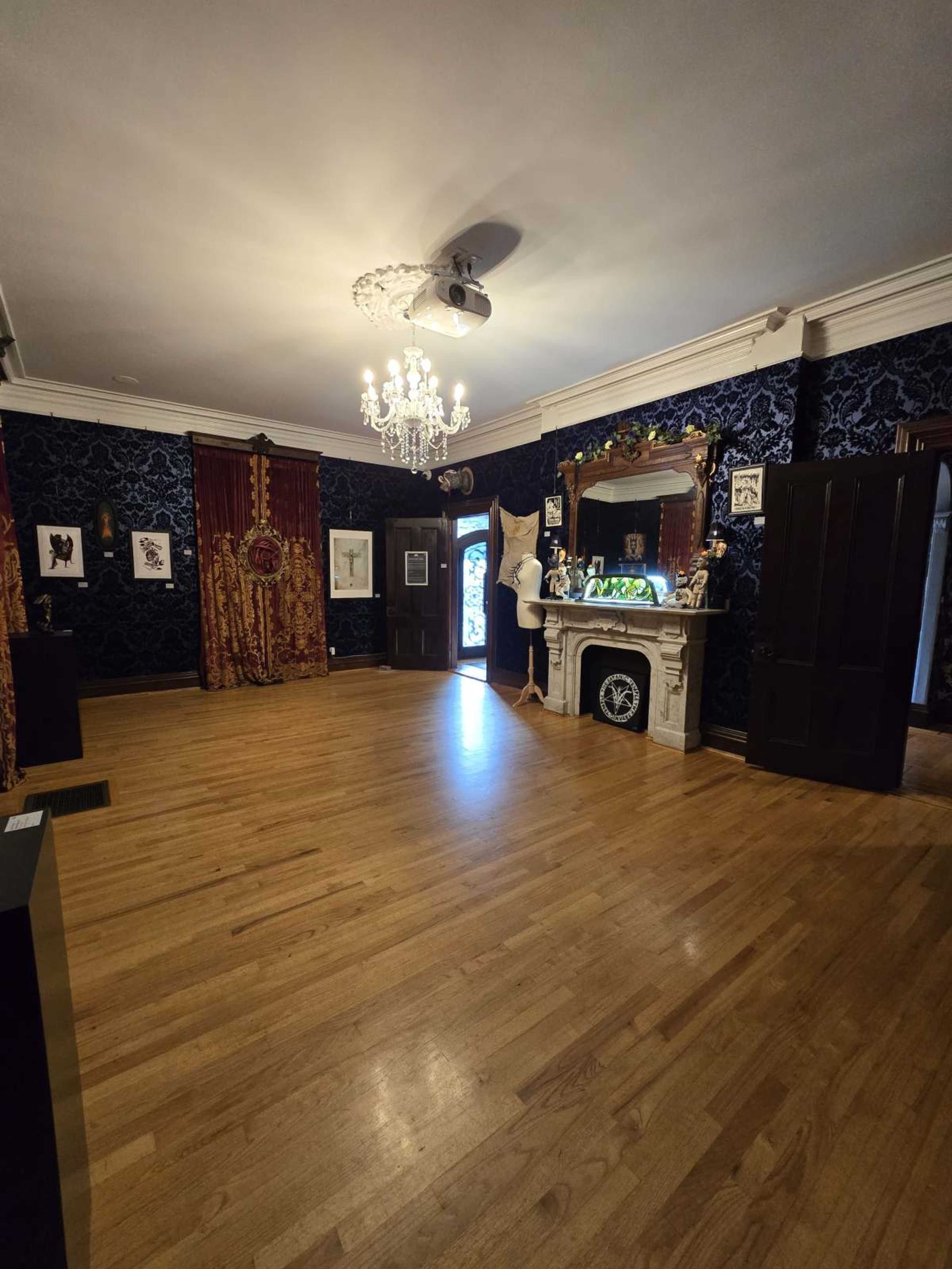 The room features polished wooden floors, dark patterned wallpaper, a chandelier, and a fireplace with decorative elements, along with an open door leading outside.