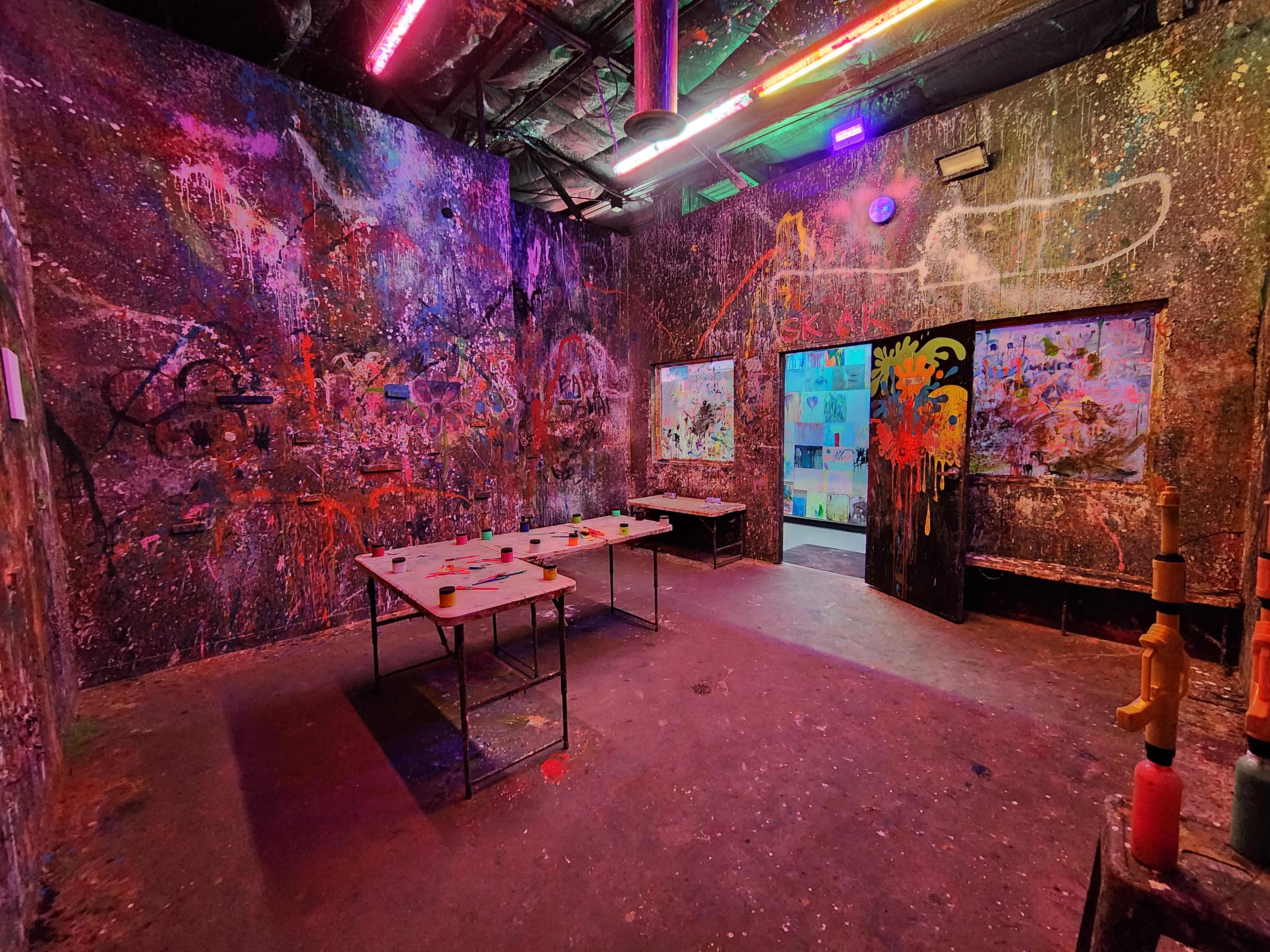The room features graffiti-covered walls and colorful neon lights, with a table set up in the center and two doors leading to other areas.
