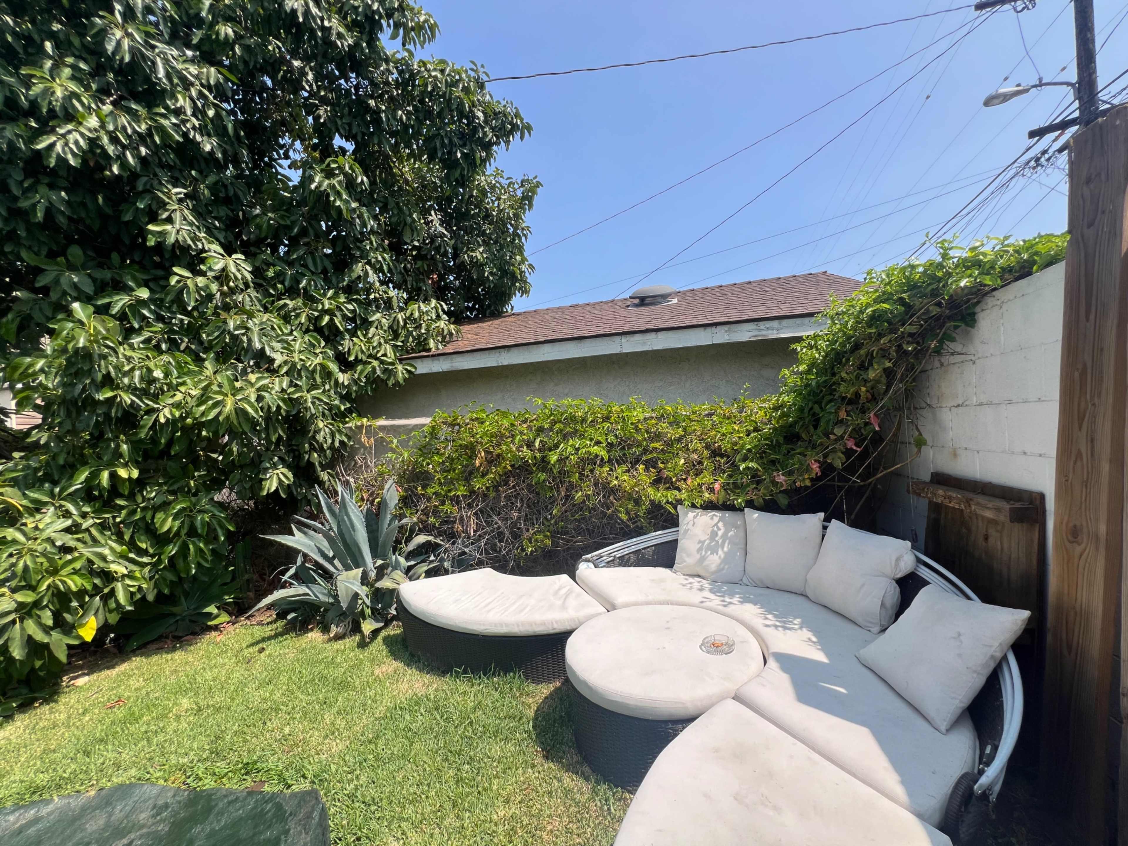 A circular outdoor seating area with cushioned furniture is located on green grass beside a flowering shrub and a tall tree.
