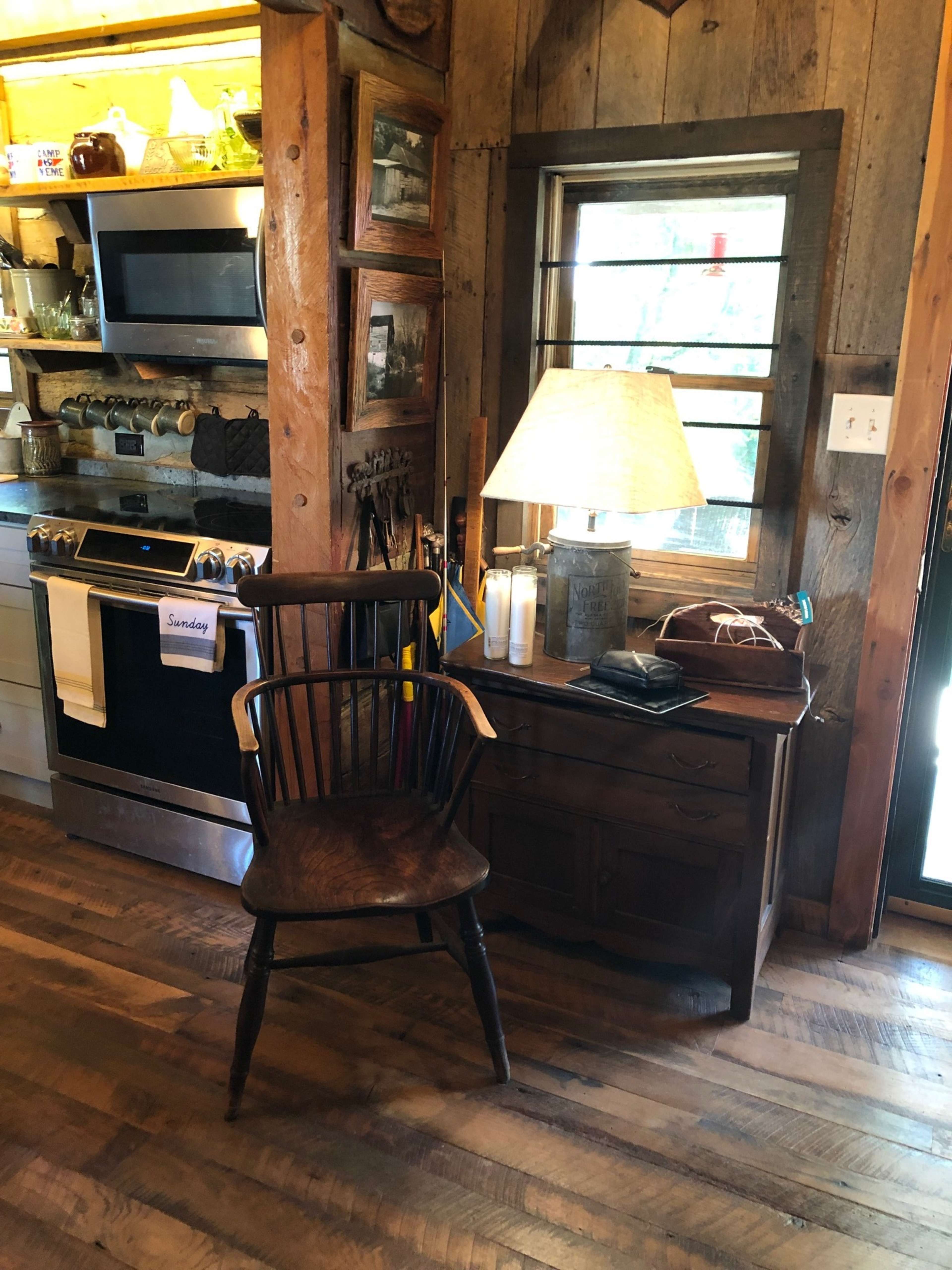 A rustic kitchen with wooden walls, a wooden chair next to a small table, and a lamp illuminating the space.