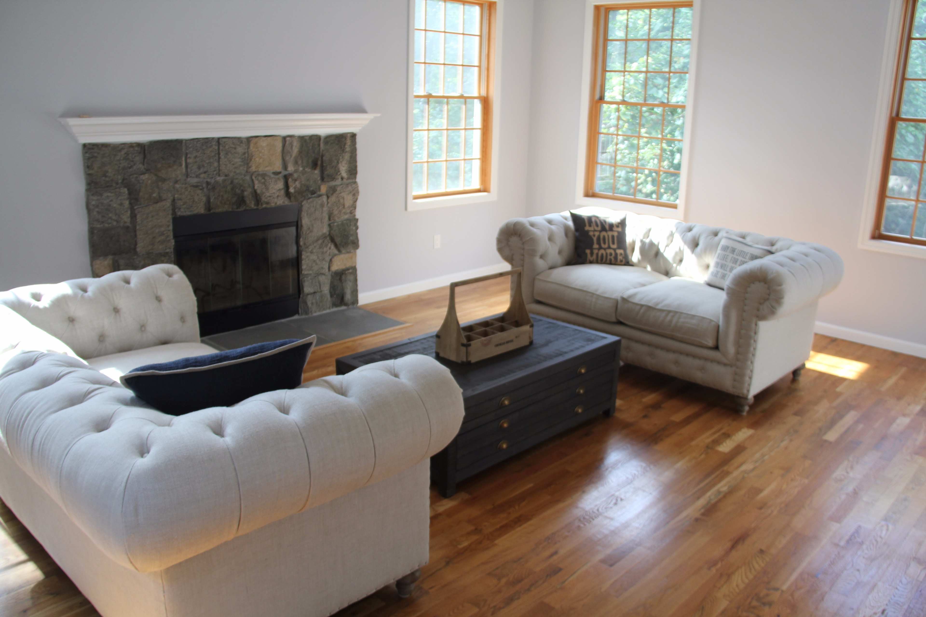 A bright living room features two tufted sofas facing a wooden coffee table, with a stone fireplace and large windows allowing natural light.
