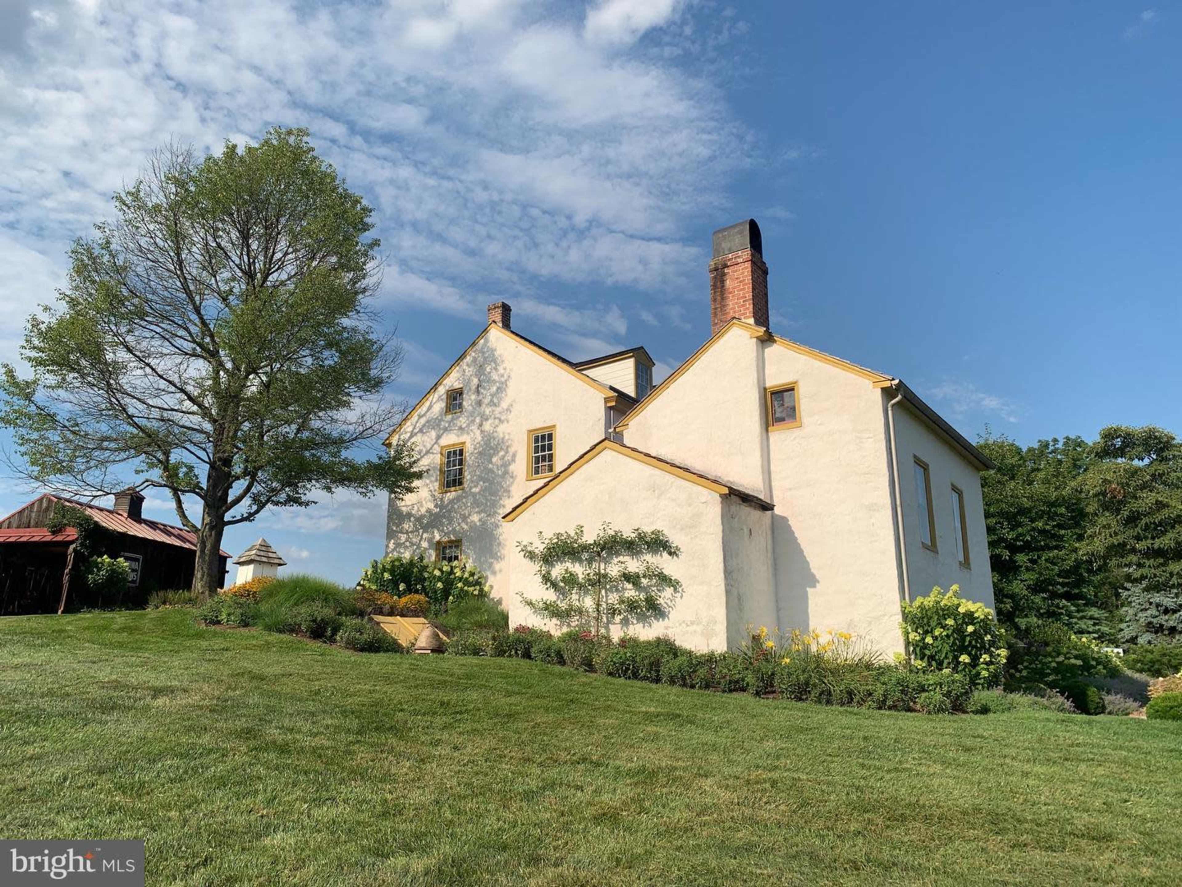 A two-story house with a light-colored exterior and yellow trim, set in a grassy area with trees and shrubs nearby.