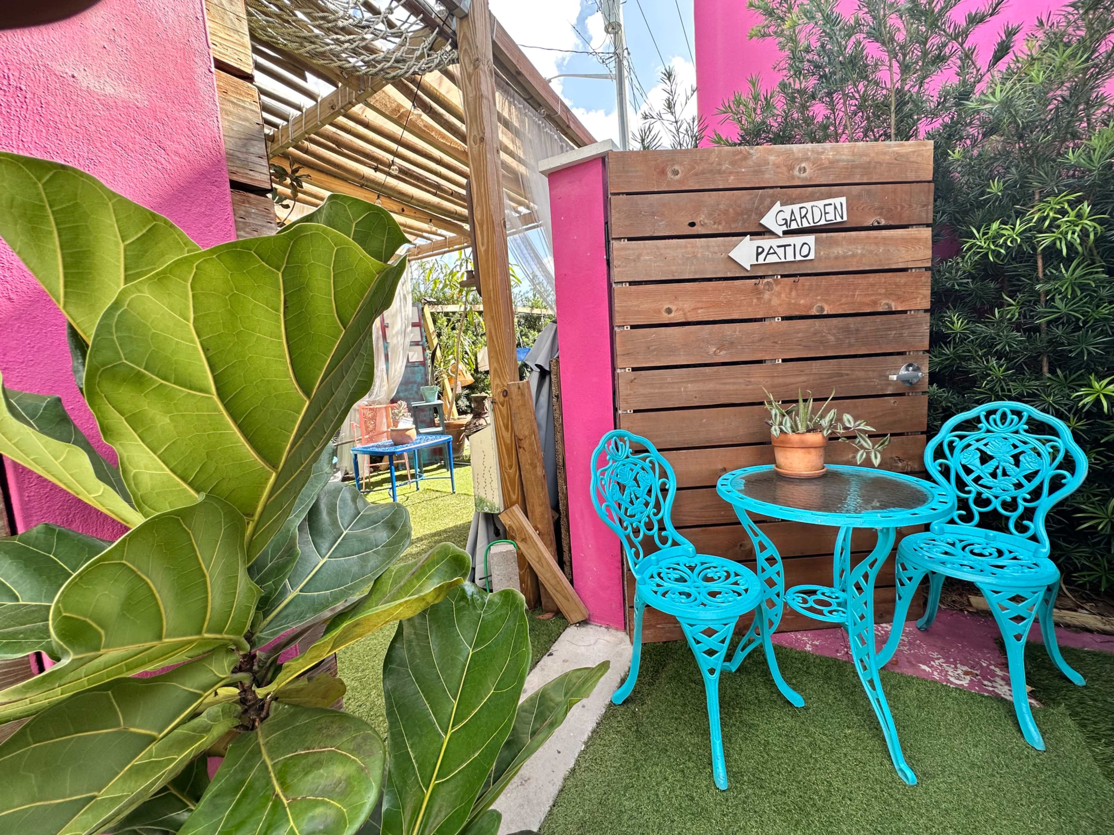 A small garden patio features a round glass table surrounded by two turquoise metal chairs and a potted plant, set against a vibrant pink wall.