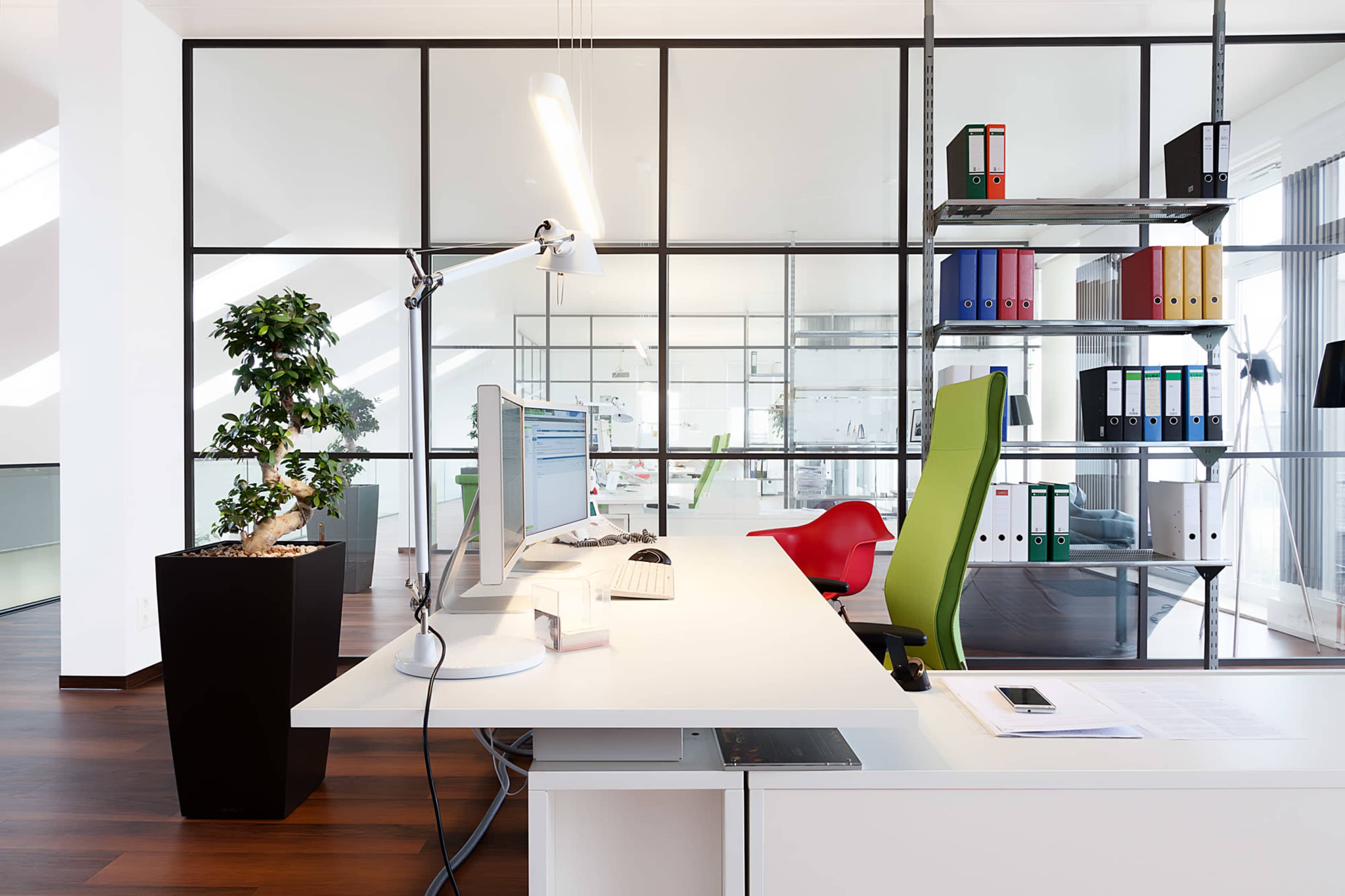 A modern office features a desk with two computer monitors, a green office chair, and a plant in a black pot, alongside shelves filled with colorful binders.