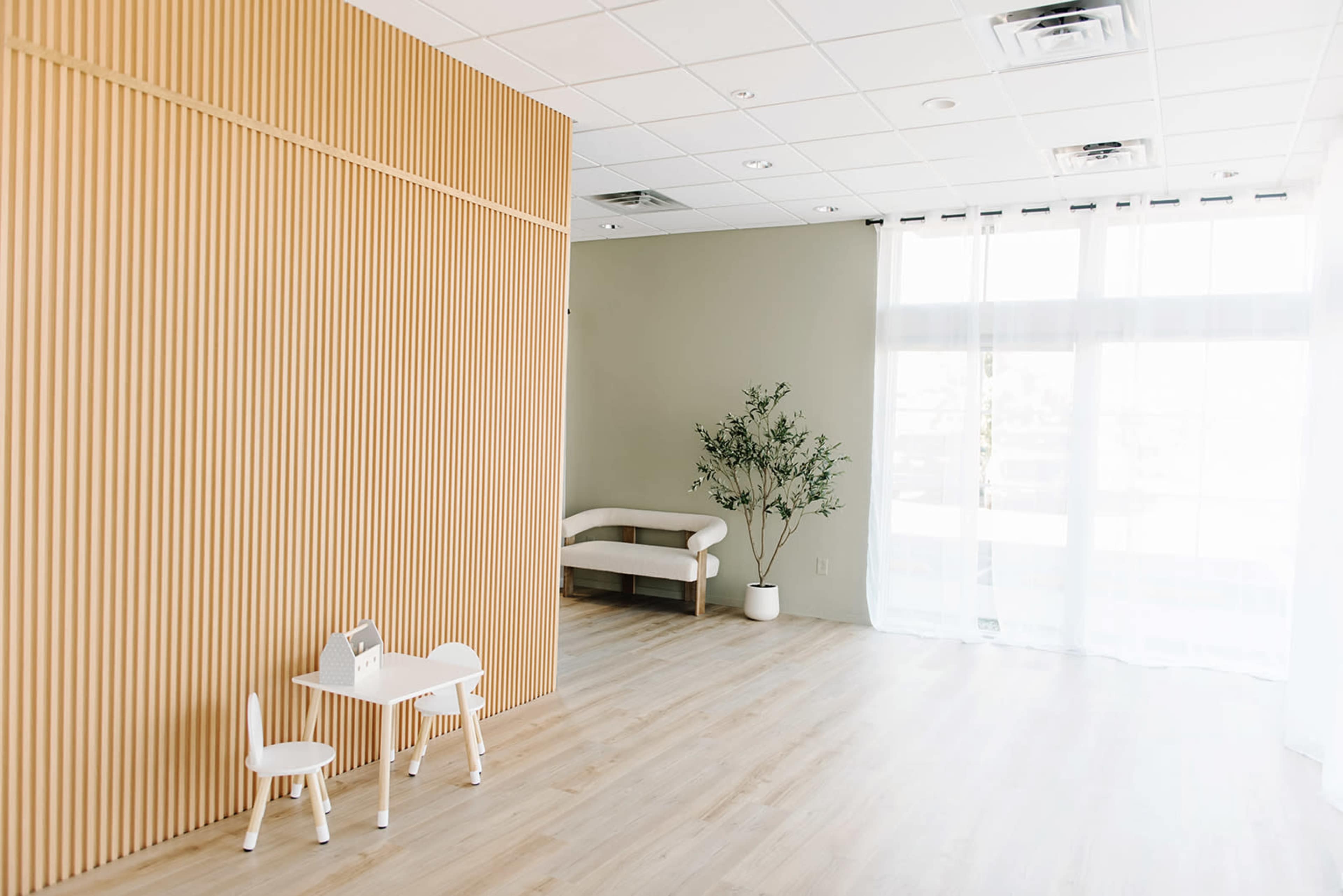 The room features a wooden-paneled wall, a small table with two chairs, a couch, and a potted plant beside large windows covered with sheer curtains.